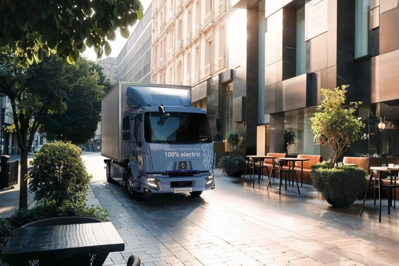 An electric delivery truck parked on a city sidewalk next to a restaurant outdoor seating area with tables, chairs, and potted plants, with sunlight streaming from the right.