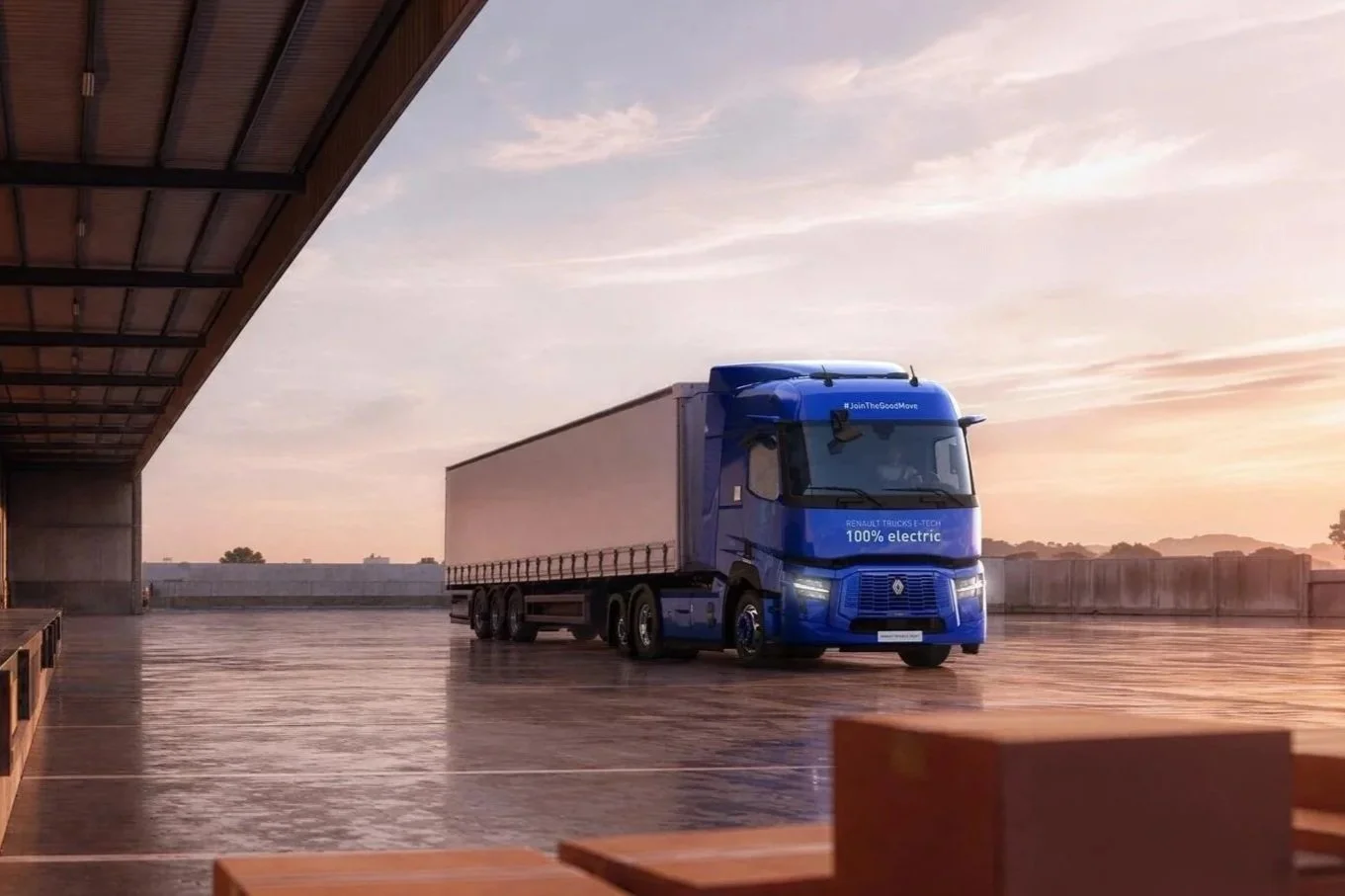 A blue electric truck parked inside a warehouse with a concrete floor and a sunset sky visible in the background.