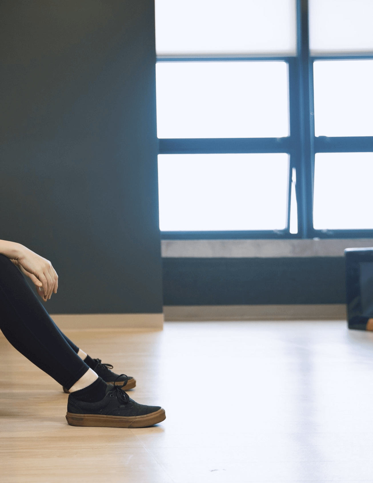 Person sitting with legs extended, wearing black sneakers and black leggings, in a room with wooden floor, gray wall, and large window.