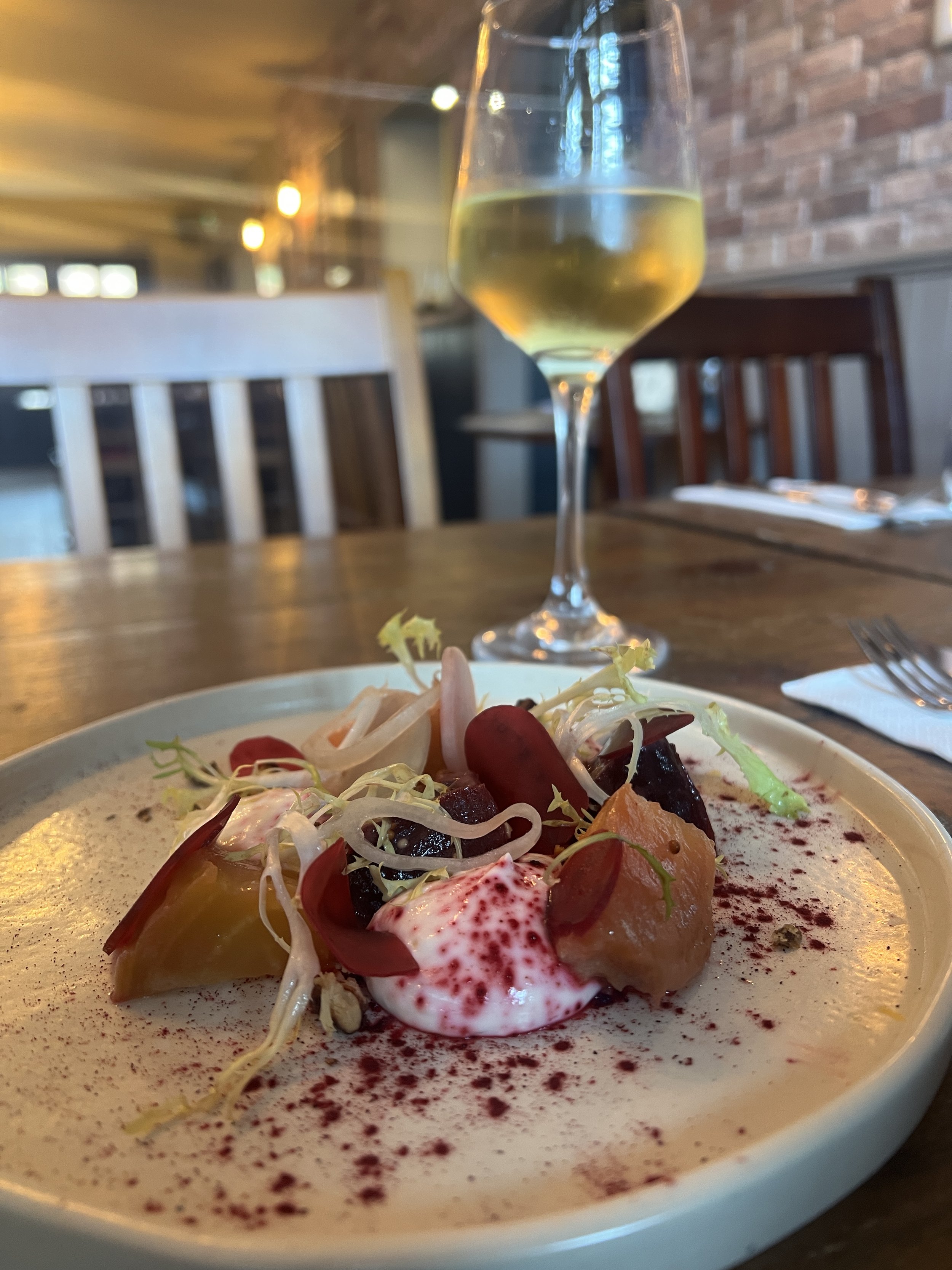 A plate of colourful salad with vegetables and creamy dressing on a wooden table, with a glass of white wine in the background inside a restaurant.