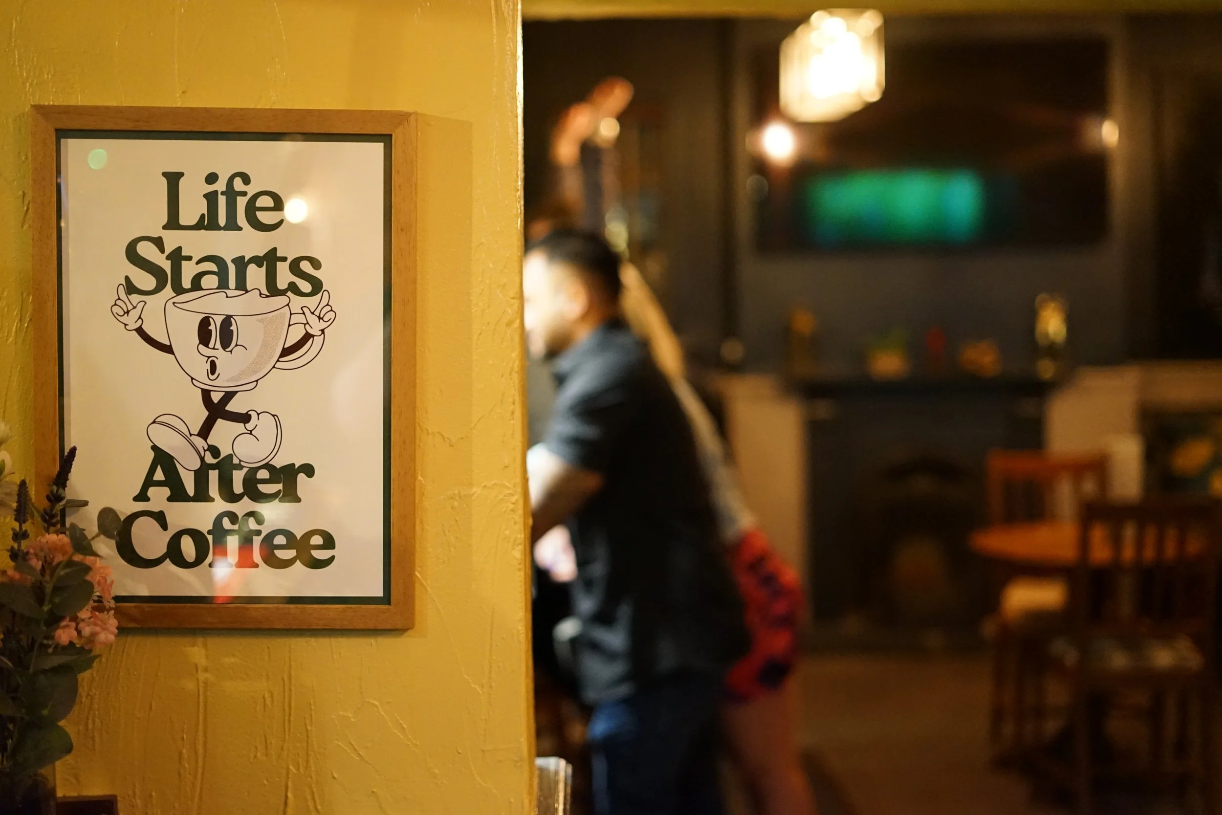 A poster on a yellow wall reading 'Life Starts After Coffee' with a cartoon coffee cup character. In the background, people are sitting and standing inside a cozy, dimly lit cafe.
