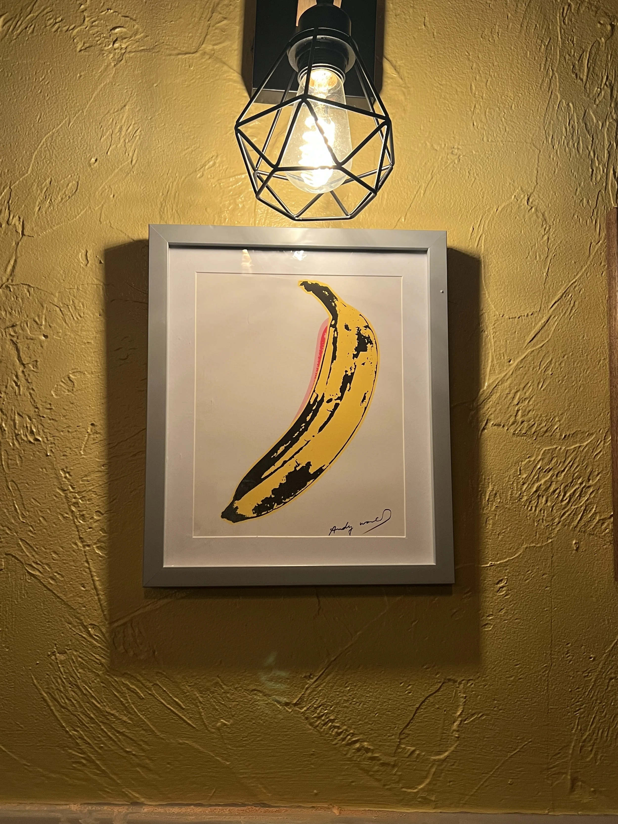 Framed artwork of a yellow banana with black spots hanging on a yellow textured wall, illuminated by a modern black metal cage pendant light.