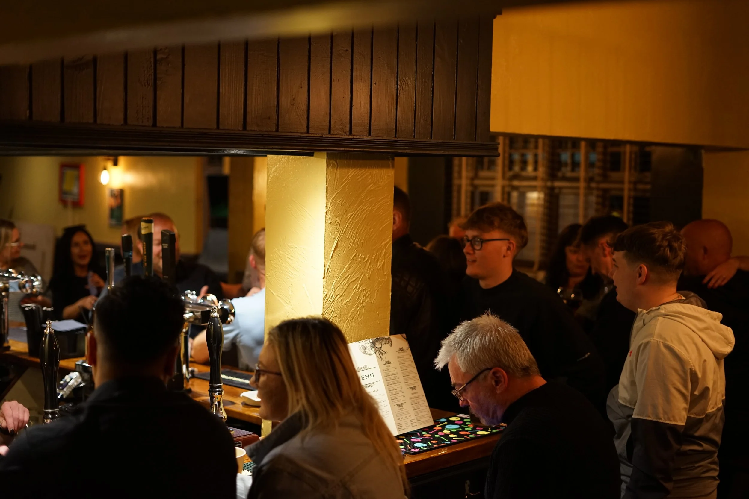 People gathered inside a bar or pub, some sitting and others standing, with beer taps visible on the counter, warm lighting, and wooden interior decor.