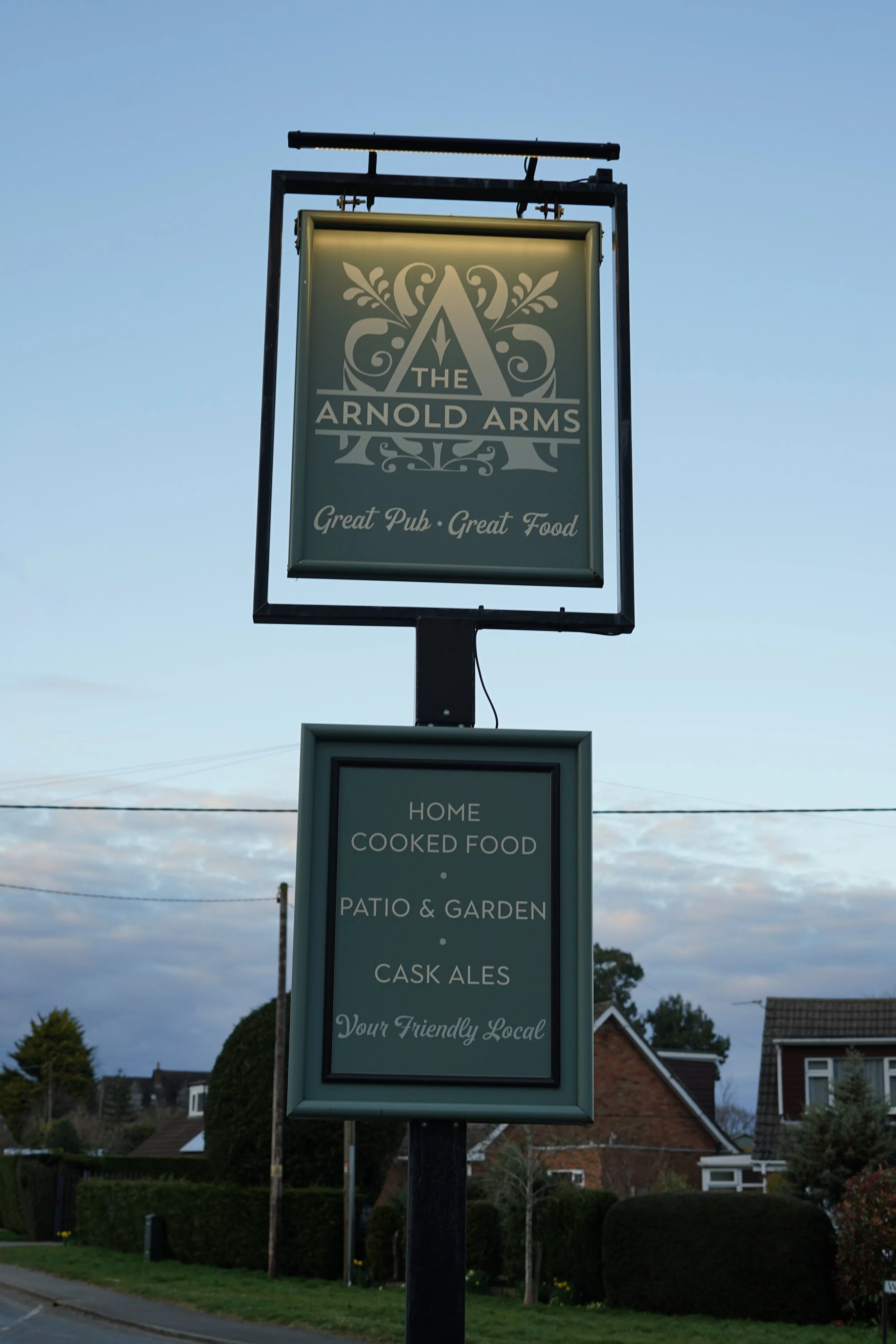 Sign for The Arnold Arms pub advertising great pub food, home cooked food, patio and garden, and cask ales, located in a suburban area with houses and trees.