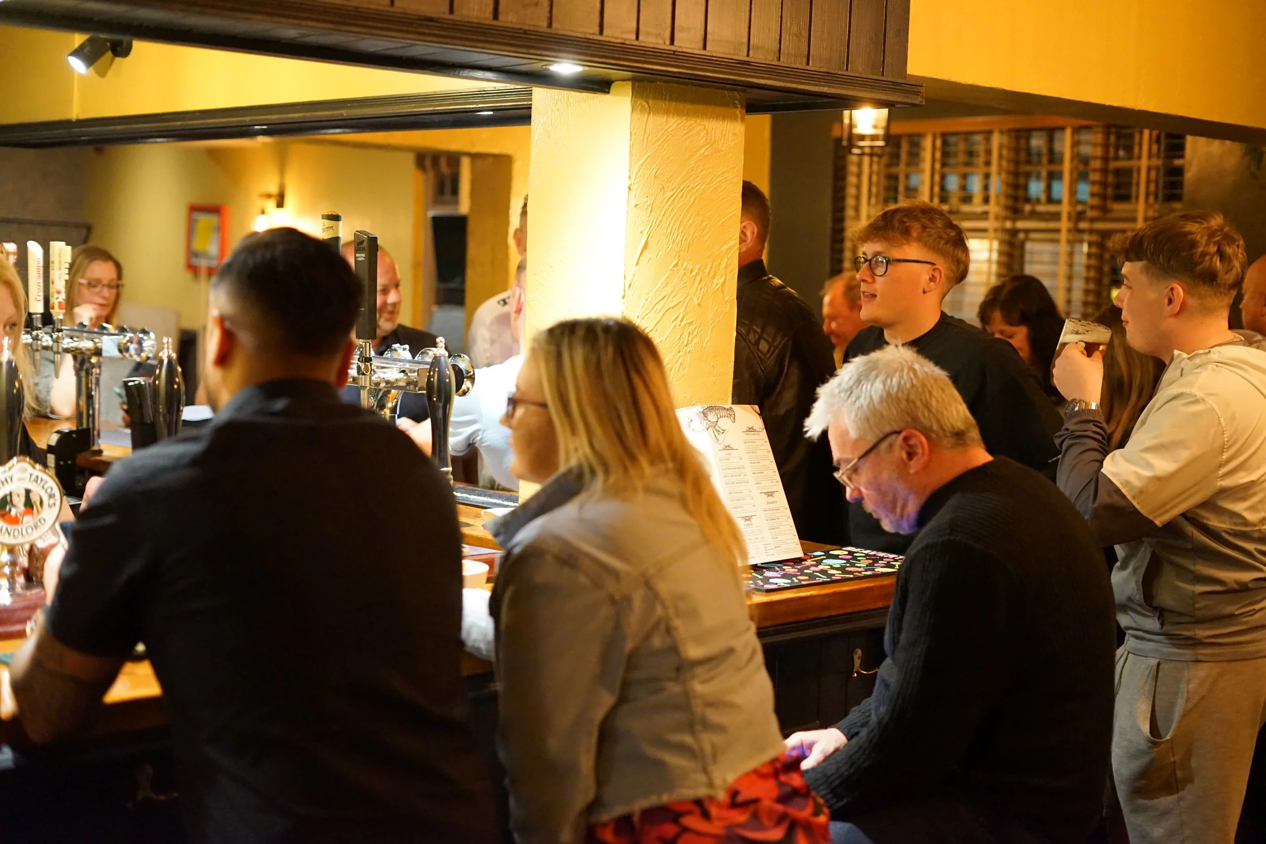People sitting and standing at a bar in a dimly lit pub, with some holding drinks and engaging in conversation.