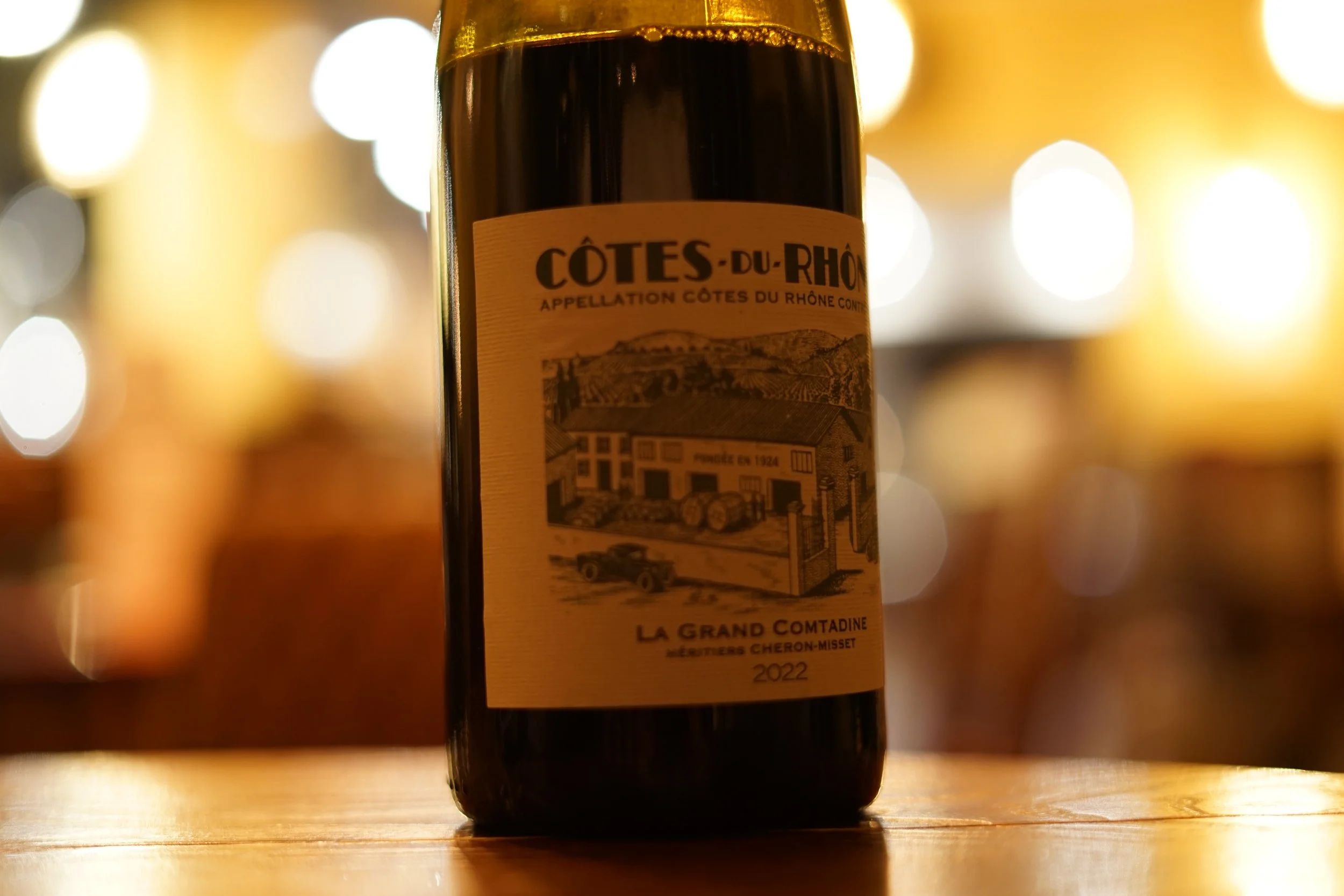 A bottle of Côtes-du-Rhône wine with a label showing a landscape and a building, placed on a wooden surface with blurred lights in the background.