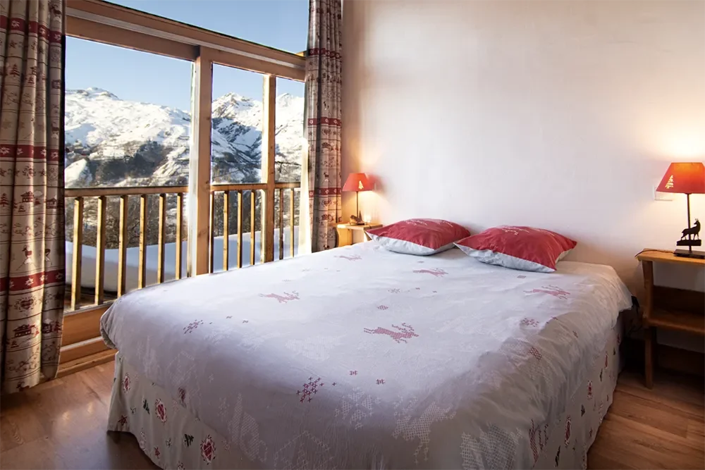 Hotel bedroom with a large bed covered in white and red bedding, two red pillows, and matching red bedside lamps. Large window showing snow-covered mountains and trees outside, with curtains on either side.