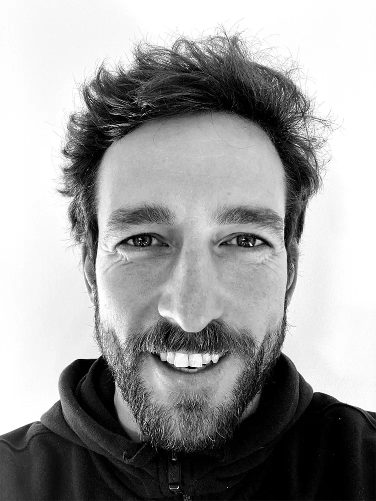 Close-up of a young man with a beard and tousled hair, smiling at the camera in black and white.