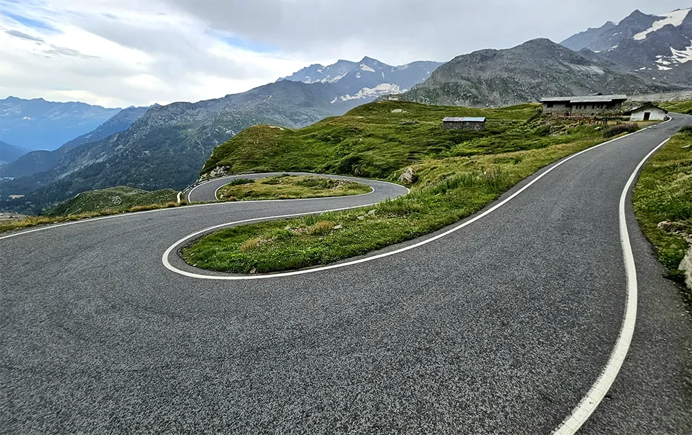 Winding mountain road with sharp curves, surrounded by green grass and rocky terrain, mountains in the background under cloudy sky.
