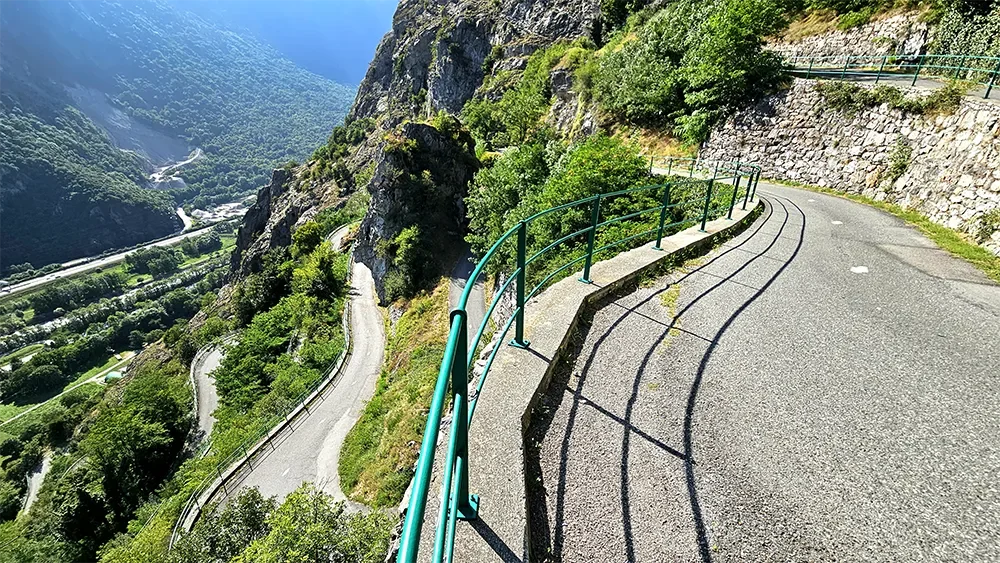A winding mountain road with a green metal guardrail on the edge of a rocky cliff, overlooking a valley with lush greenery and river below.