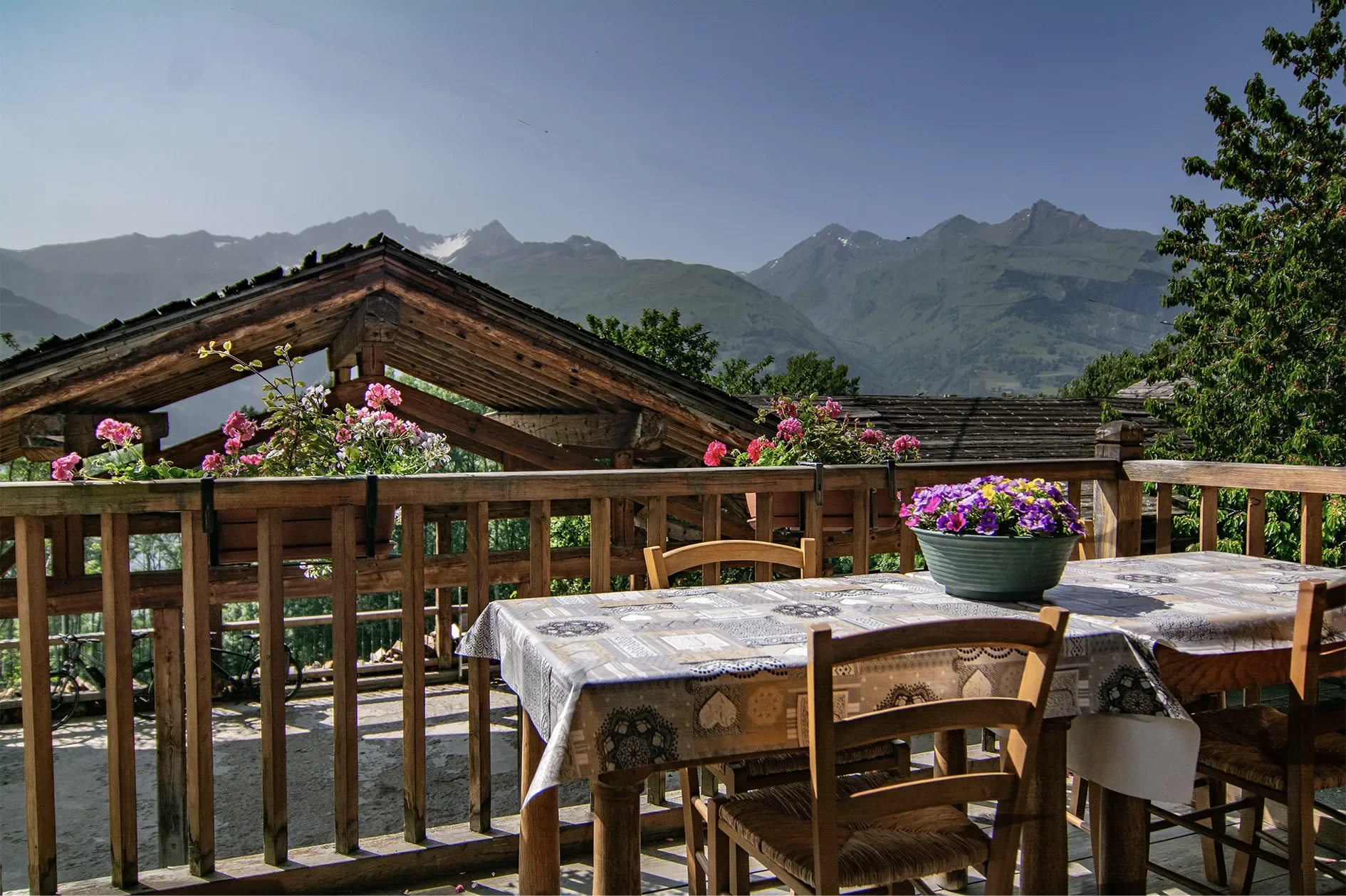 A wooden outdoor balcony with a table covered by a patterned tablecloth and decorated with a flower pot and potted flowers, with mountain scenery and trees in the background.