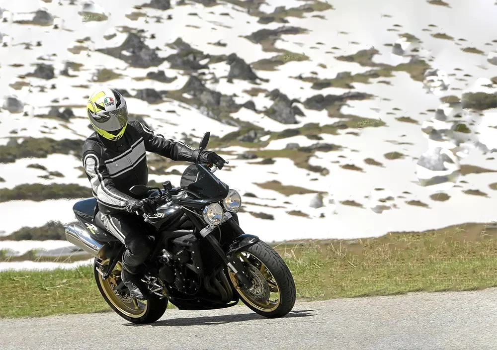 Motorcycle rider in black riding gear with yellow helmet on winding road in a mountainous landscape with snow patches.