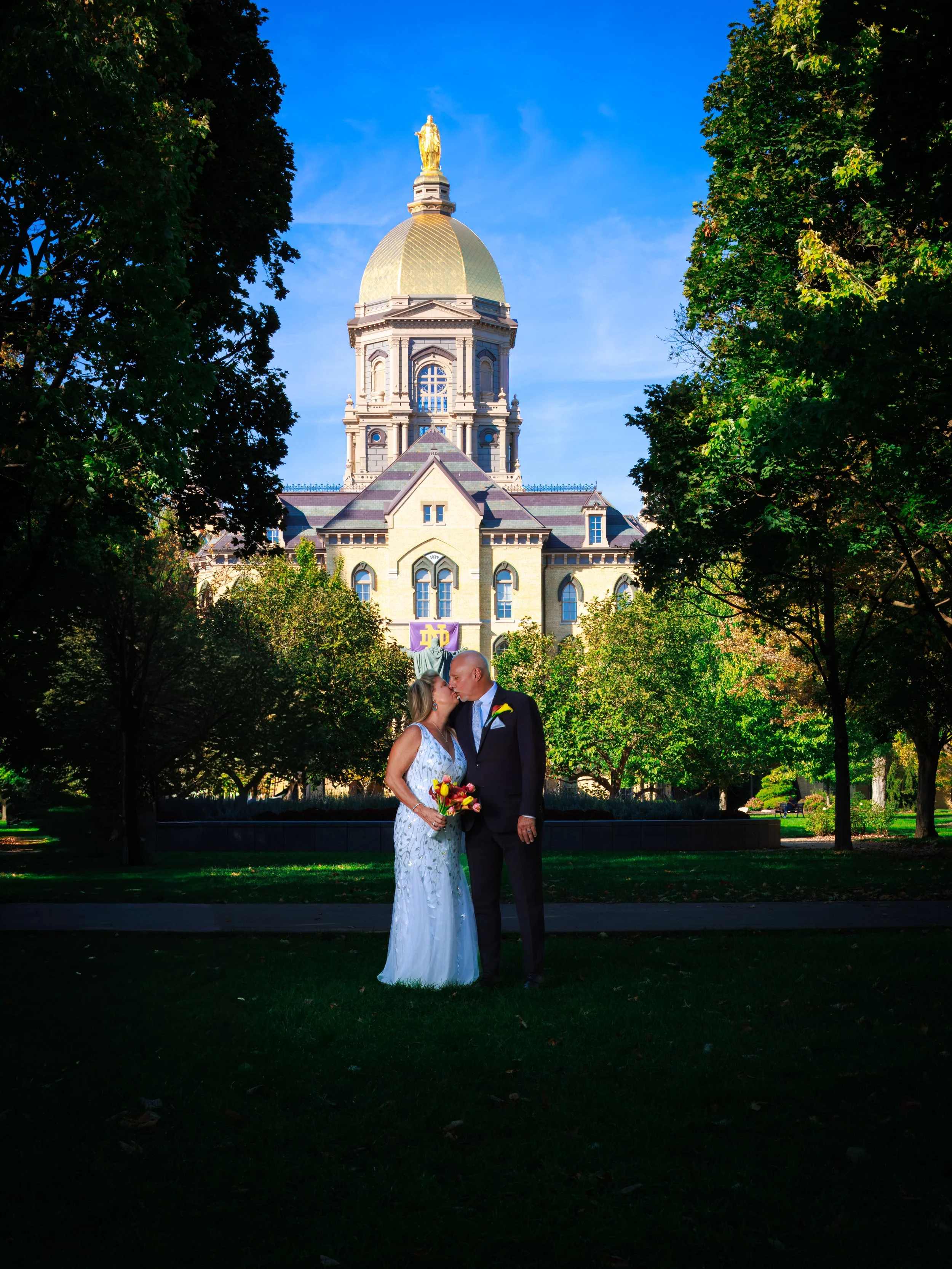A bride and groom standing in front of a large historic building with a gold dome, surrounded by trees and green grass. The bride is holding a bouquet and they are sharing a kiss.