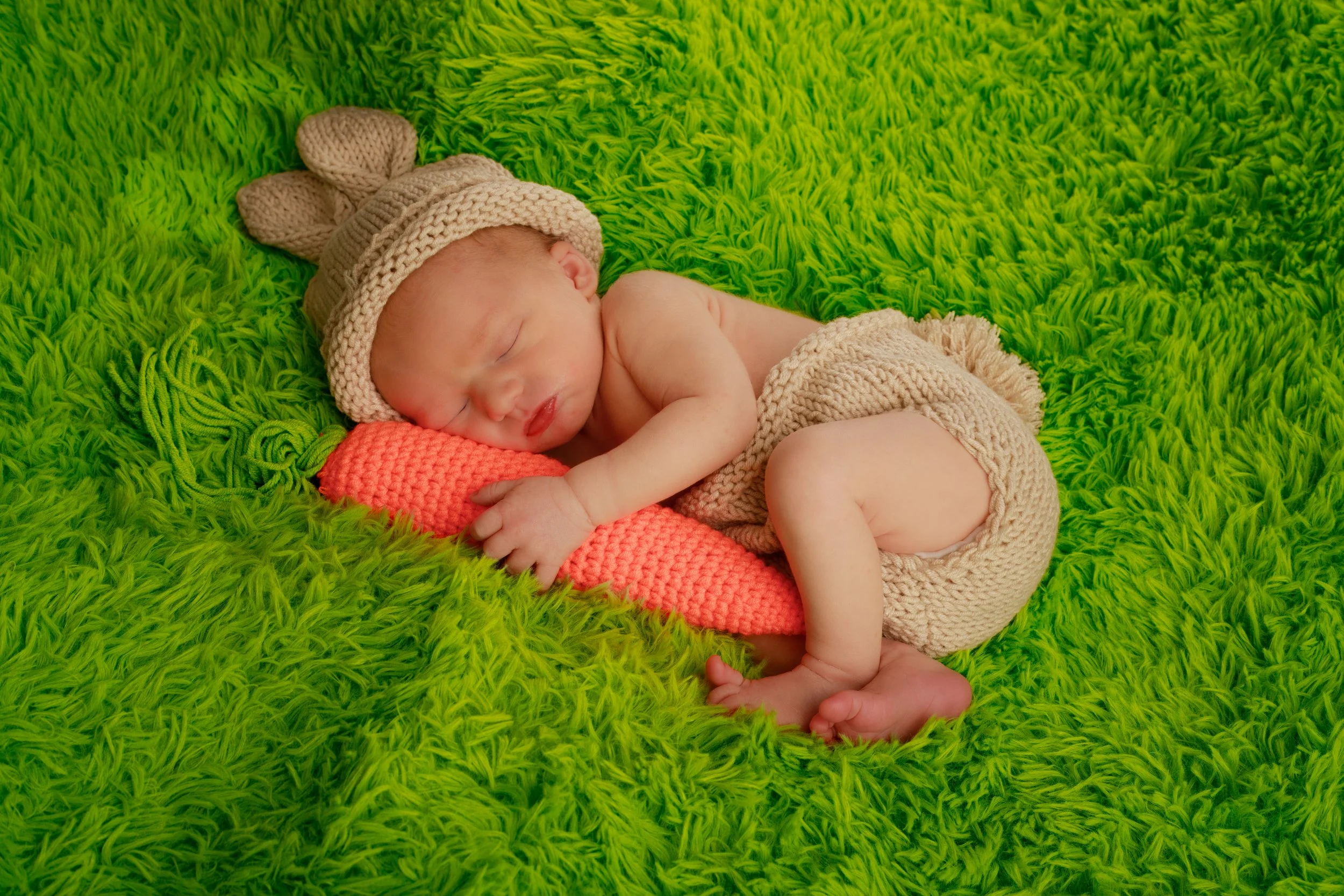 A sleeping newborn baby lying on a bright green furry blanket, wearing a knitted beige hat with bunny ears and matching knitted pants, holding a pink crocheted stuffed item.