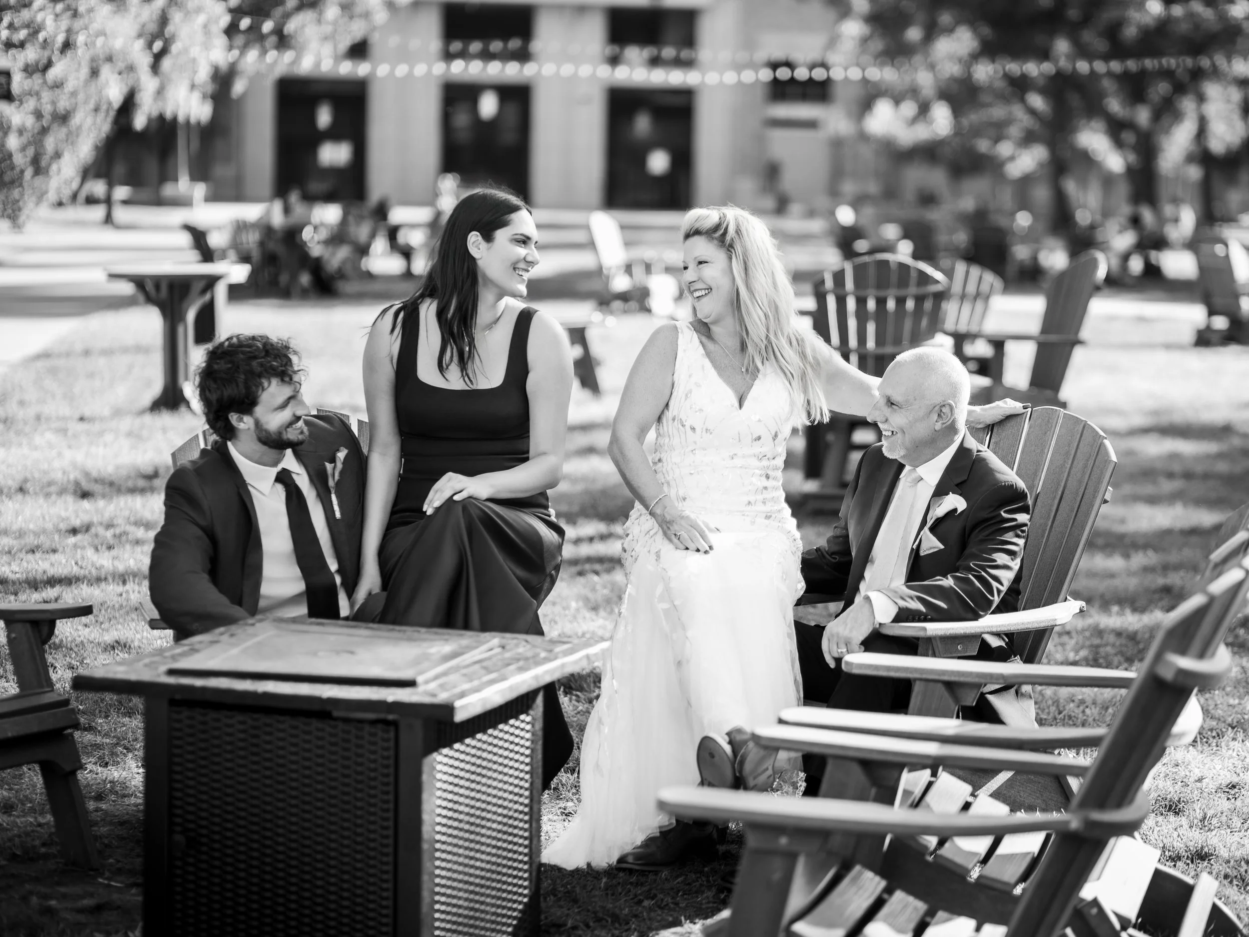 Four people, two women and two men, sit and stand in a park, smiling and talking. One woman wears a white dress, the other a black dress. One man is in a suit with a tie, the other has a beard and curly hair. Trees and park benches are in the backgro