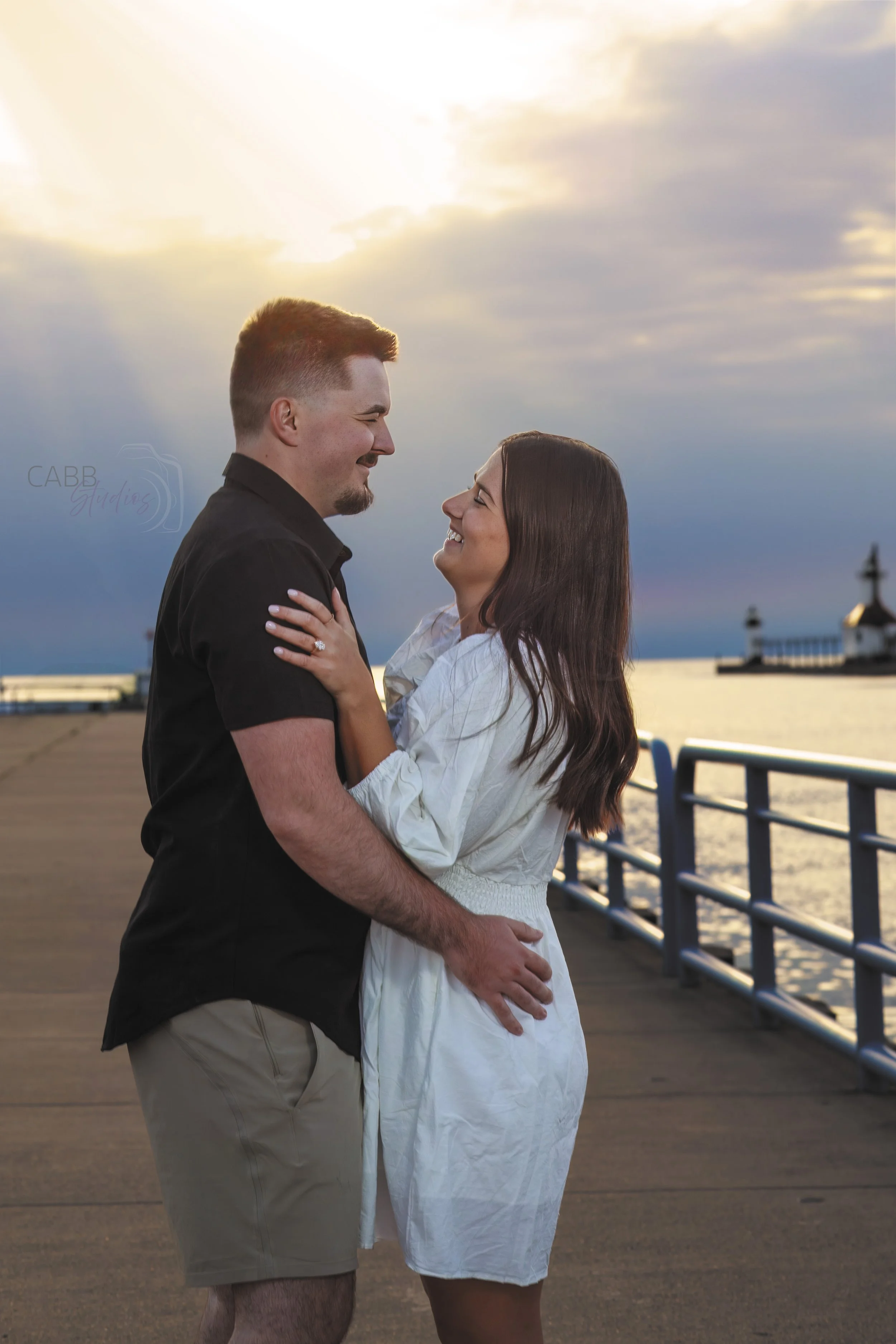 A couple smiling at each other on a pier during sunset with a lighthouse in the background.