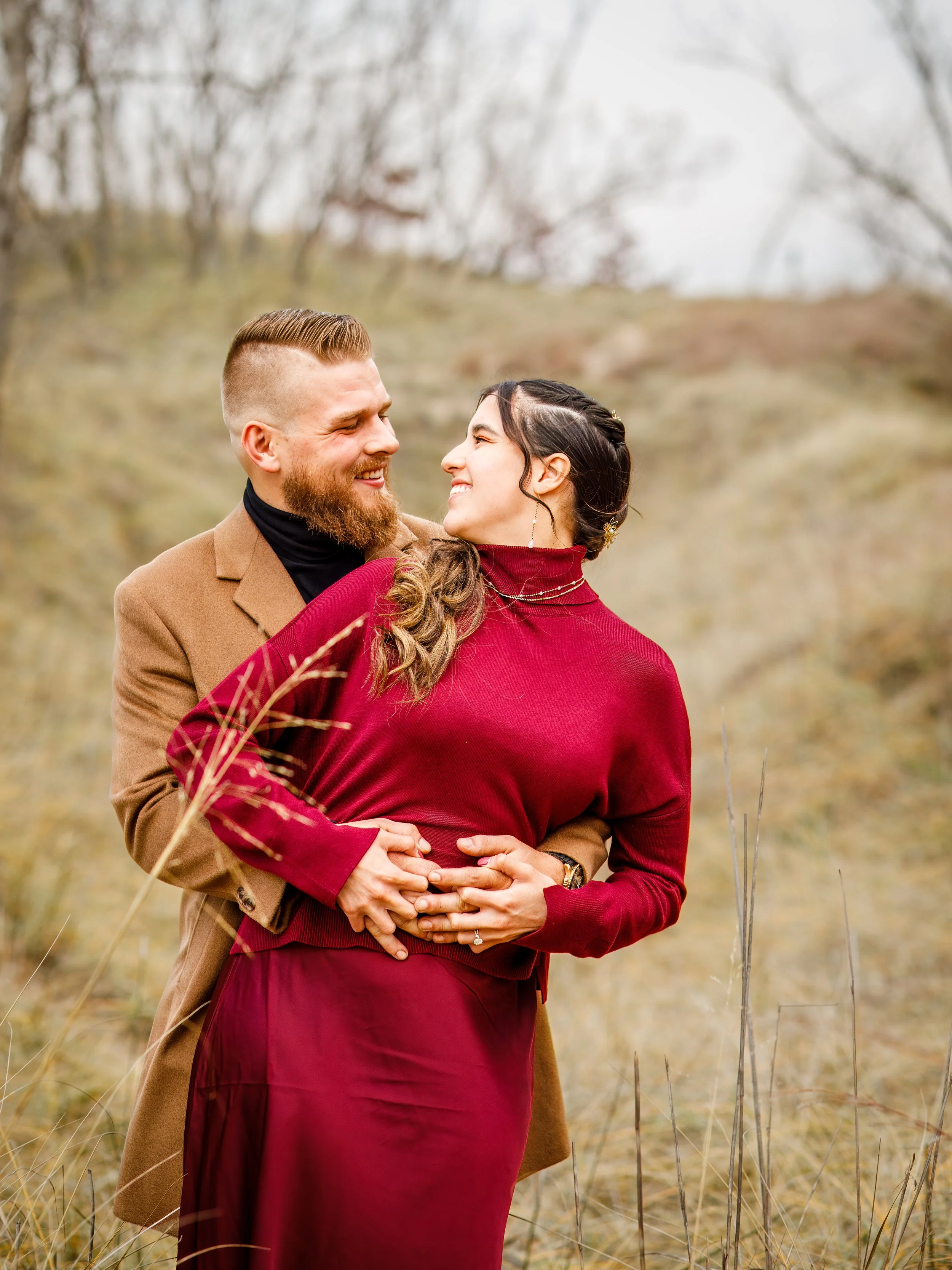 A happy couple embraces outdoors in a field with tall grass, smiling at each other.
