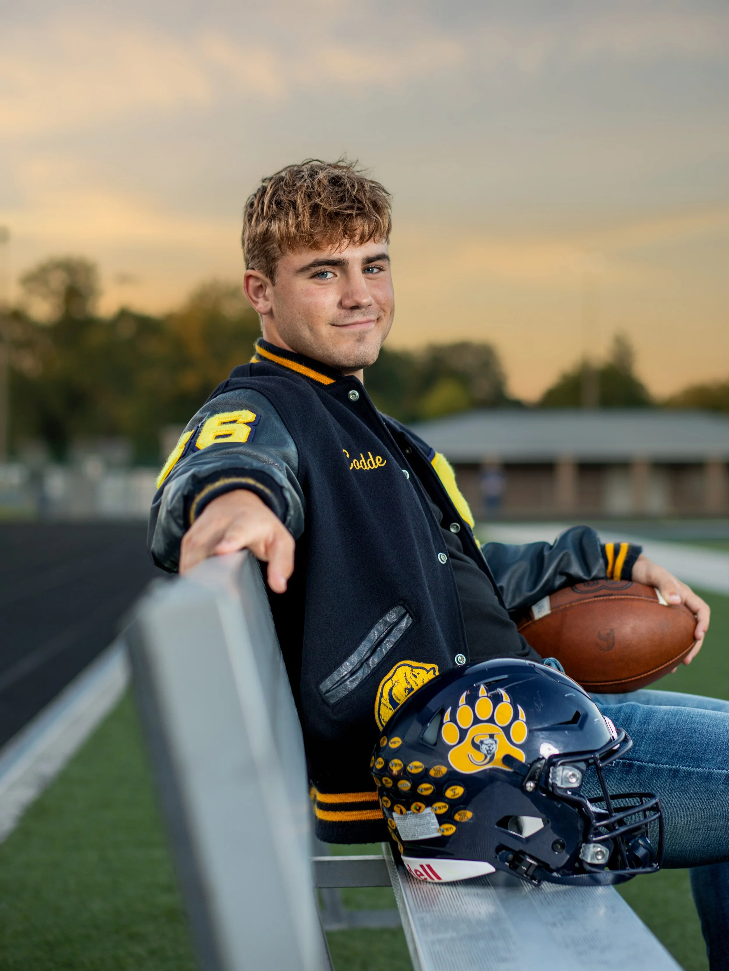 Young male football player sitting on stadium bleacher, holding a football, with a helmet on the bench next to him, at sunset.