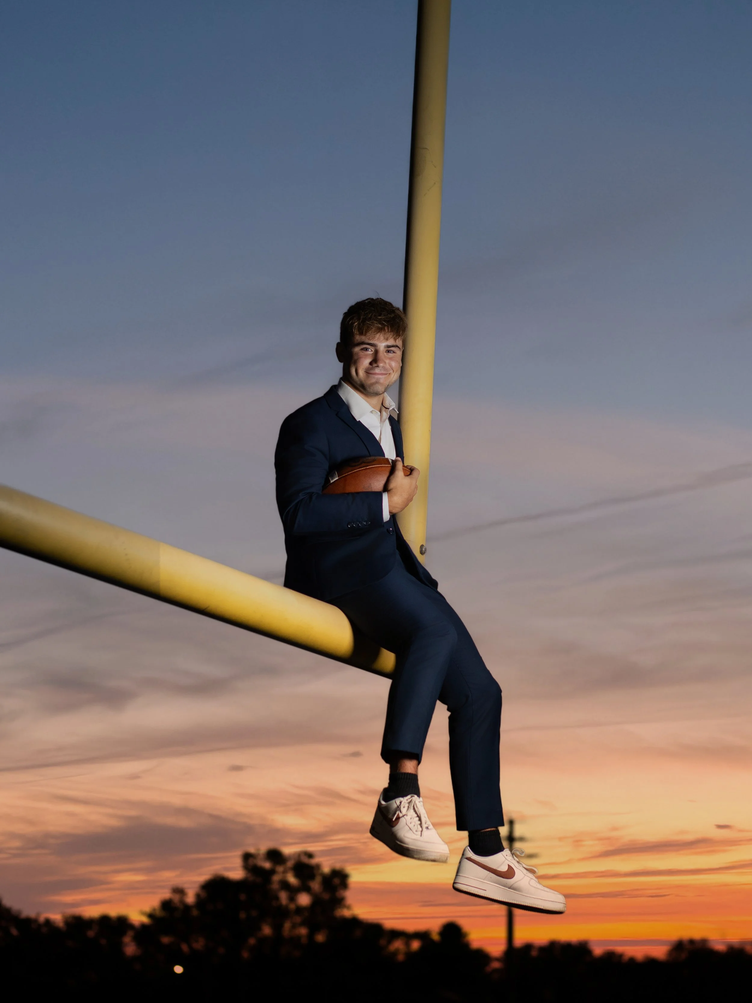 Young man in a navy suit sitting on a yellow football goal post, holding a football, during sunset with a colorful cloudy sky and silhouetted trees in the background.