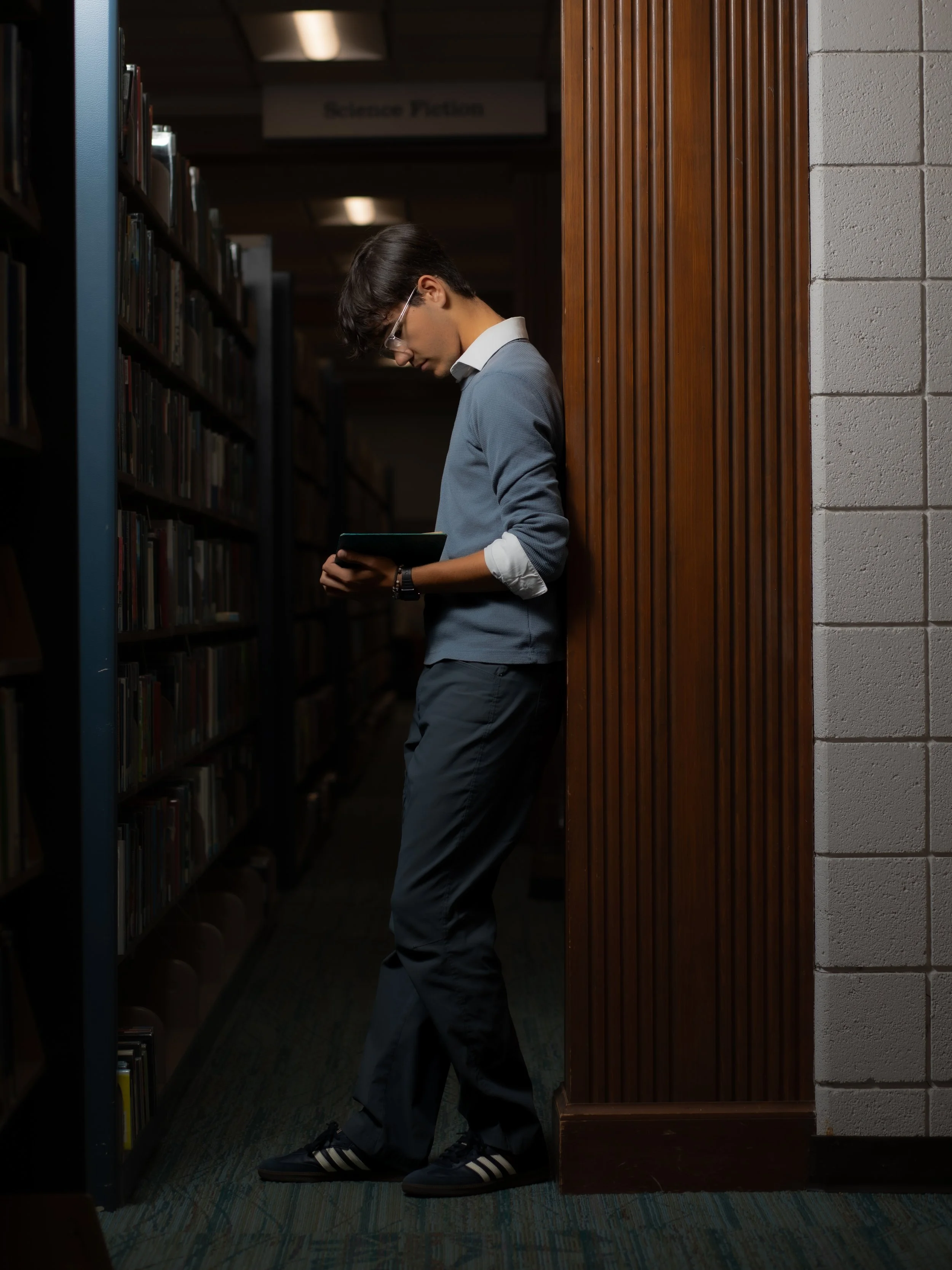 A young man in glasses, a blue sweater, and black Adidas sneakers is standing in a library, leaning against a wooden panel, reading a book.