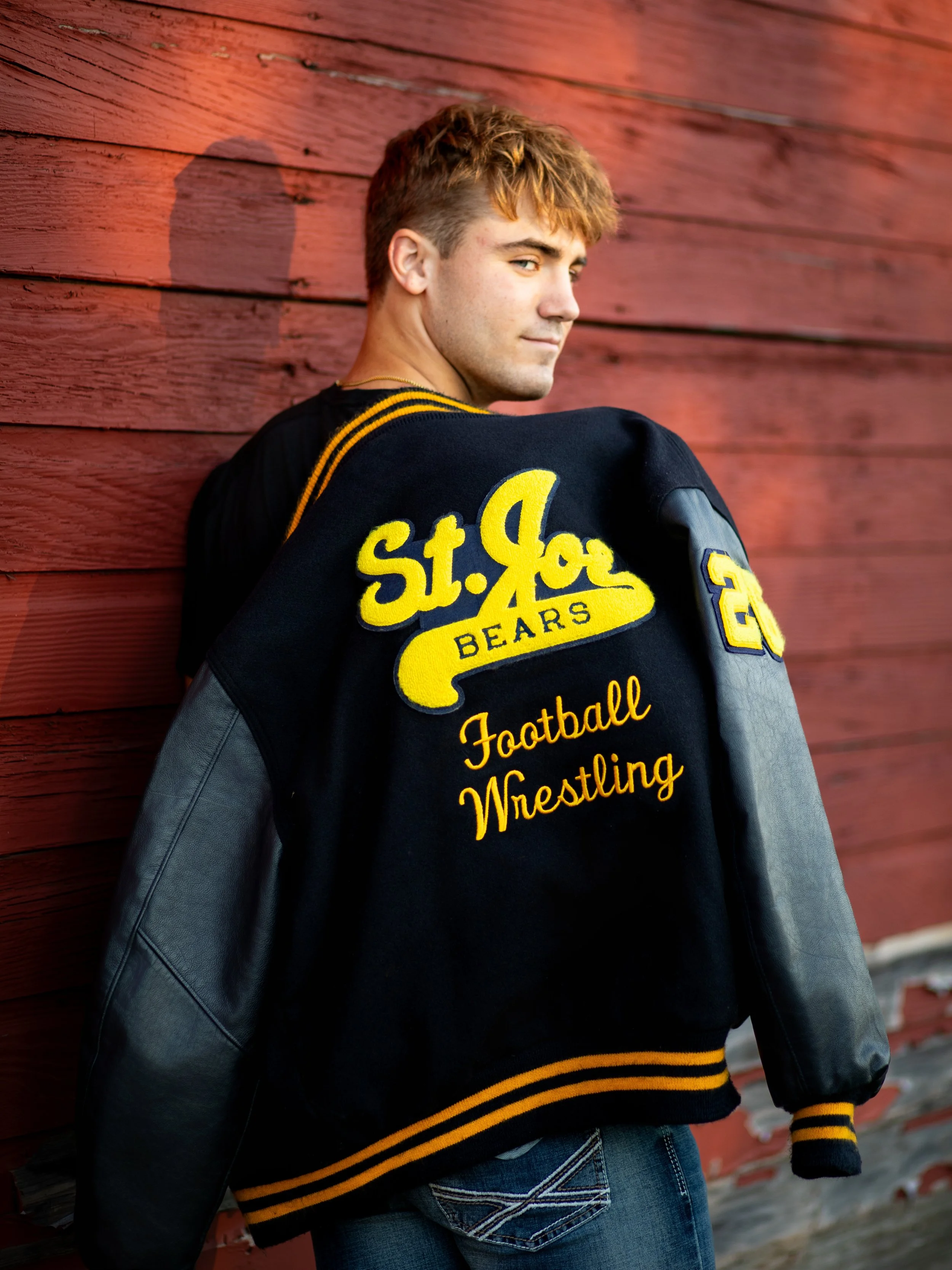 Young man leaning against a red wooden wall, wearing a black varsity jacket with yellow embroidered text and patches, looking over his shoulder.