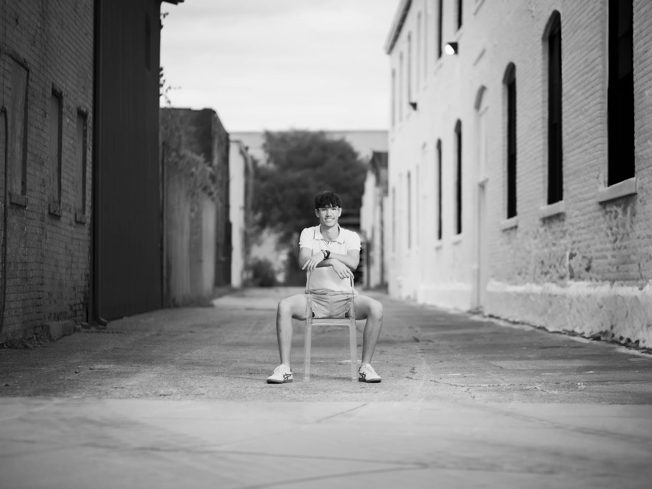 A young man sitting on a chair in an alleyway between brick buildings, smiling, in black and white.