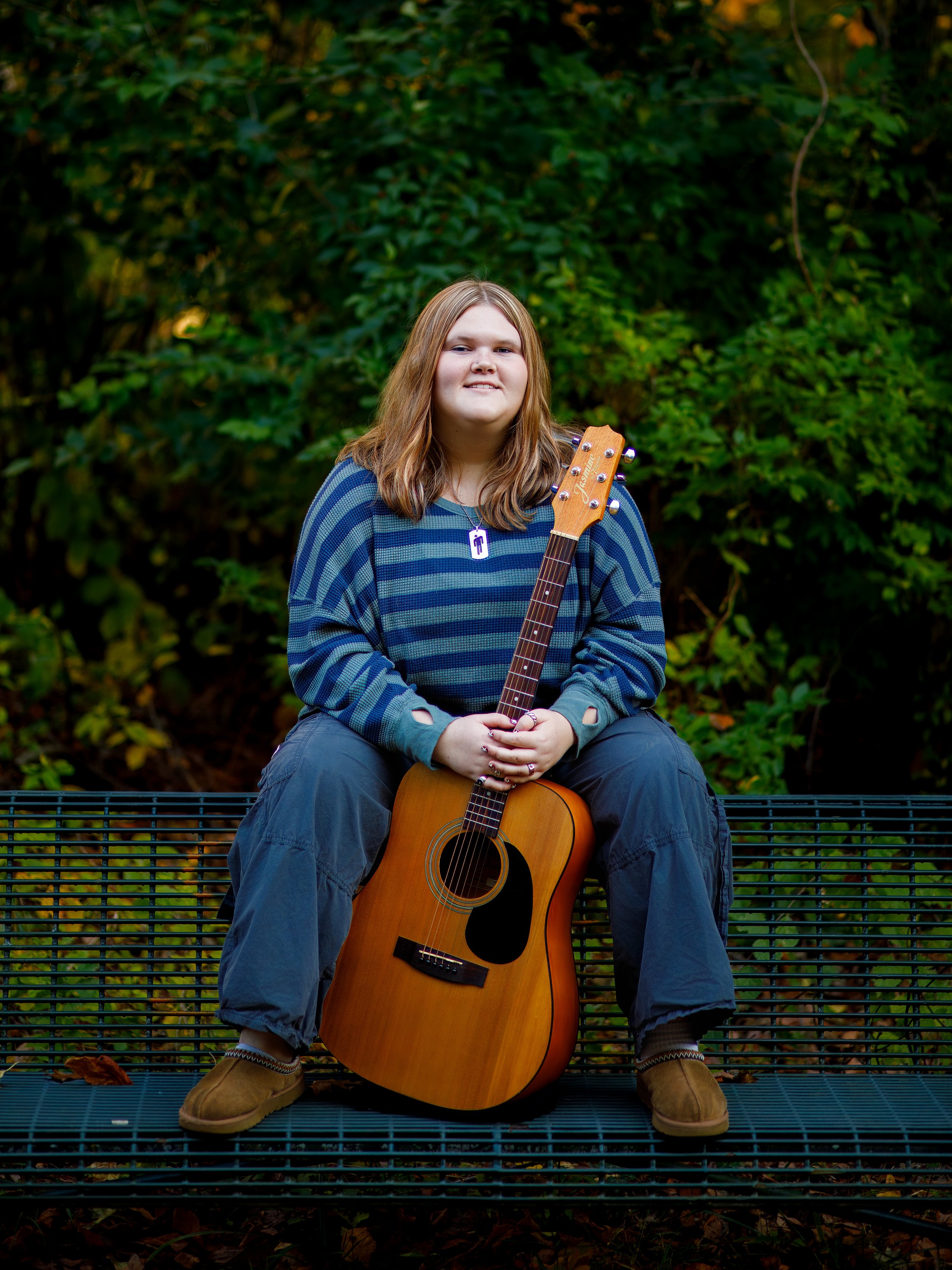 A young woman with long, reddish hair sitting on a black metal bench outdoors, holding an acoustic guitar across her lap, wearing a blue striped long sleeve shirt, dark cargo pants, and tan shoes, with green foliage in the background.