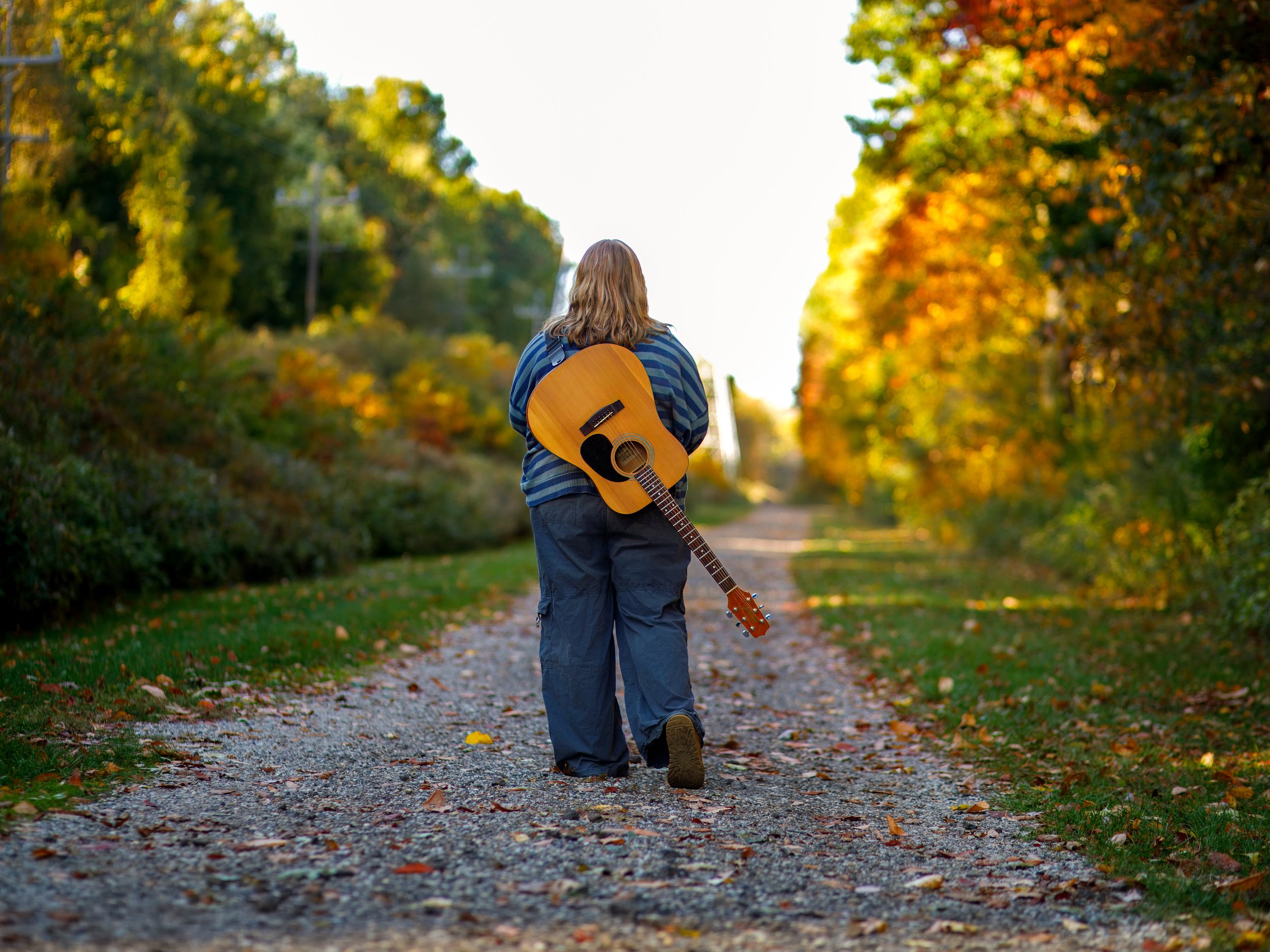 Person walking on a gravel path with a guitar on their back during fall, surrounded by colorful autumn trees.