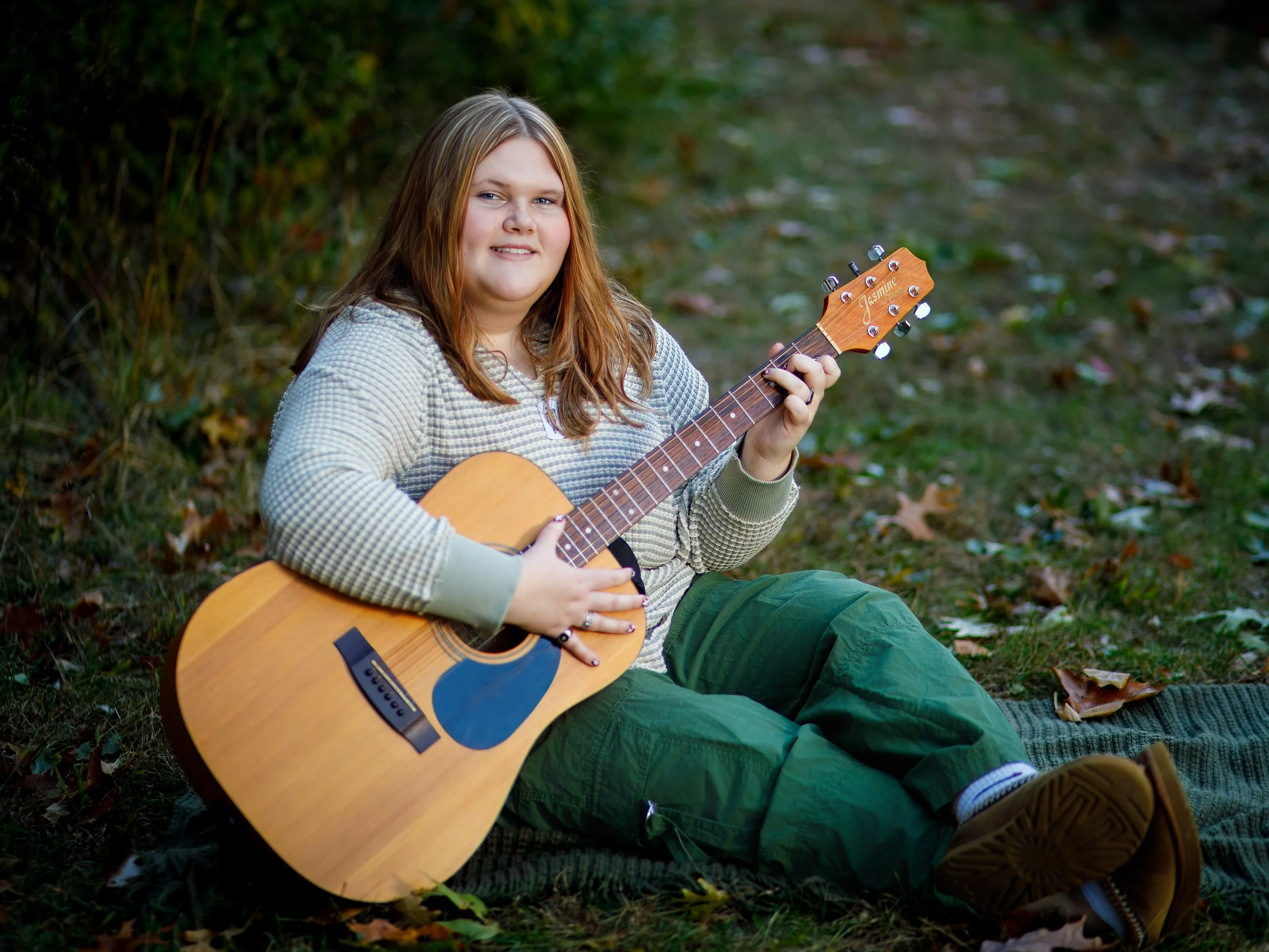 Young woman sitting on a plaid blanket outdoors, holding an acoustic guitar, surrounded by fallen leaves.