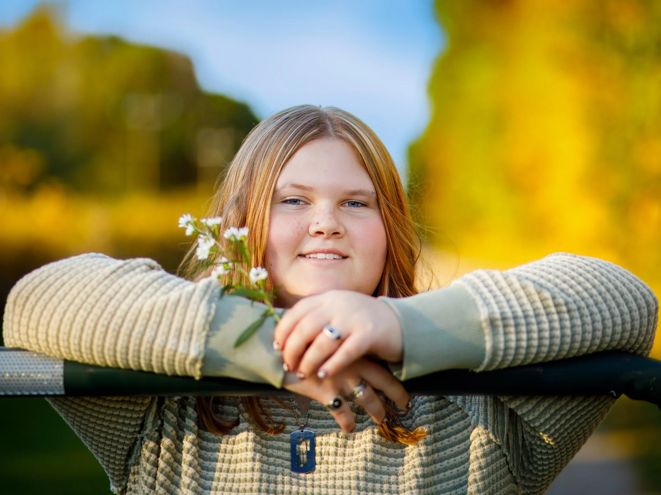 A young girl with red hair and blue eyes resting her chin on her arms on a railing, holding some small white flowers, outdoors with a blurred background of trees and a blue sky.