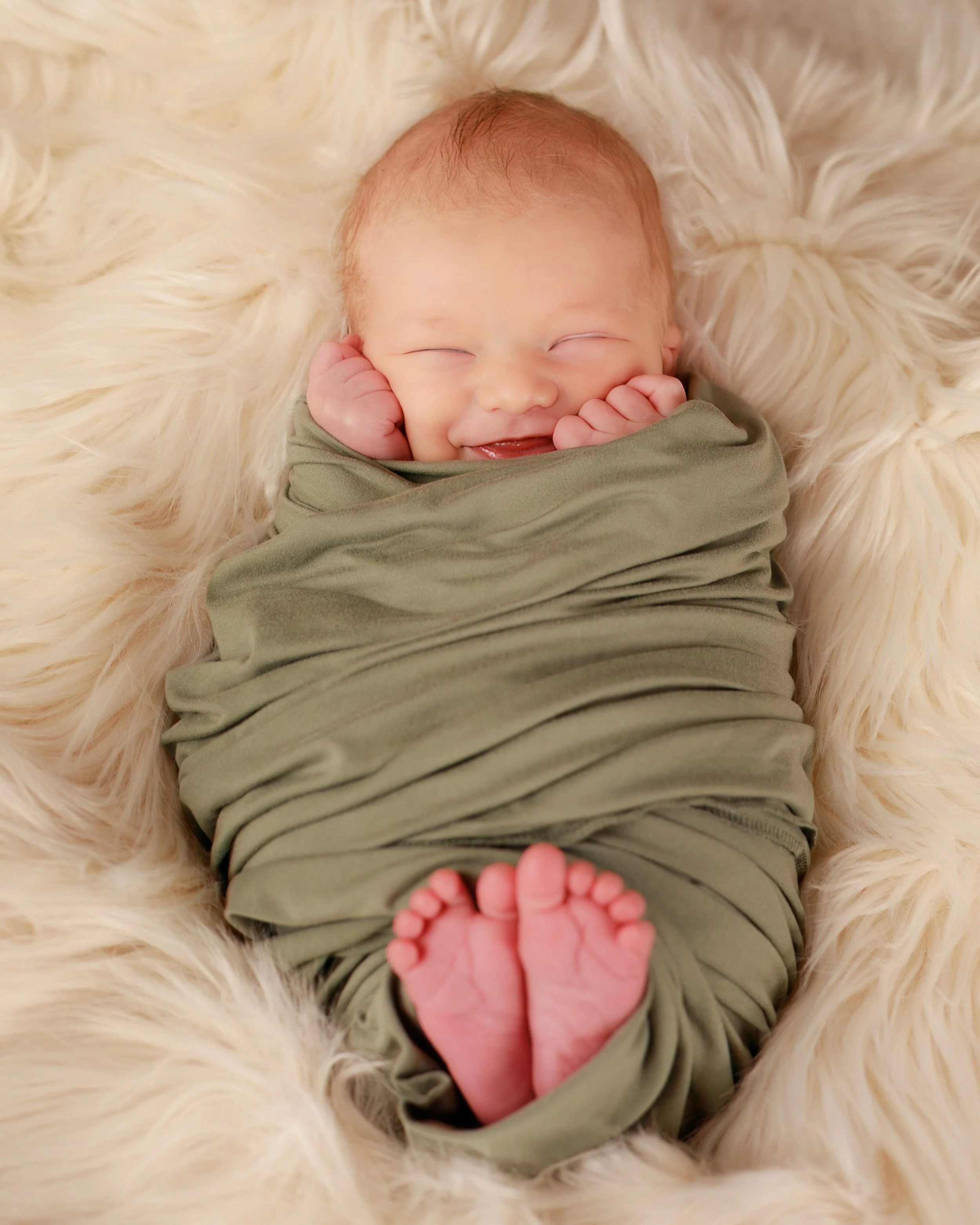 Smiling baby wrapped in green cloth, lying on a fluffy cream-colored blanket.