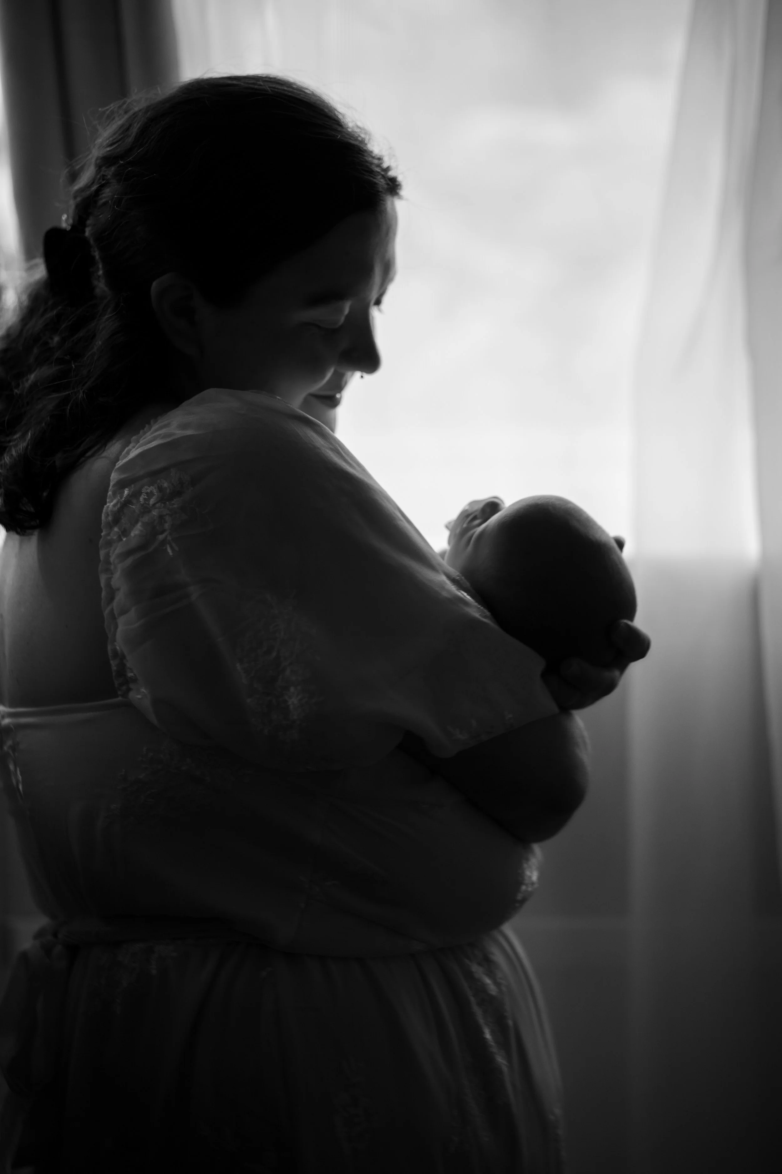 A woman holding a newborn baby, silhouetted against a bright window, smiling gently.