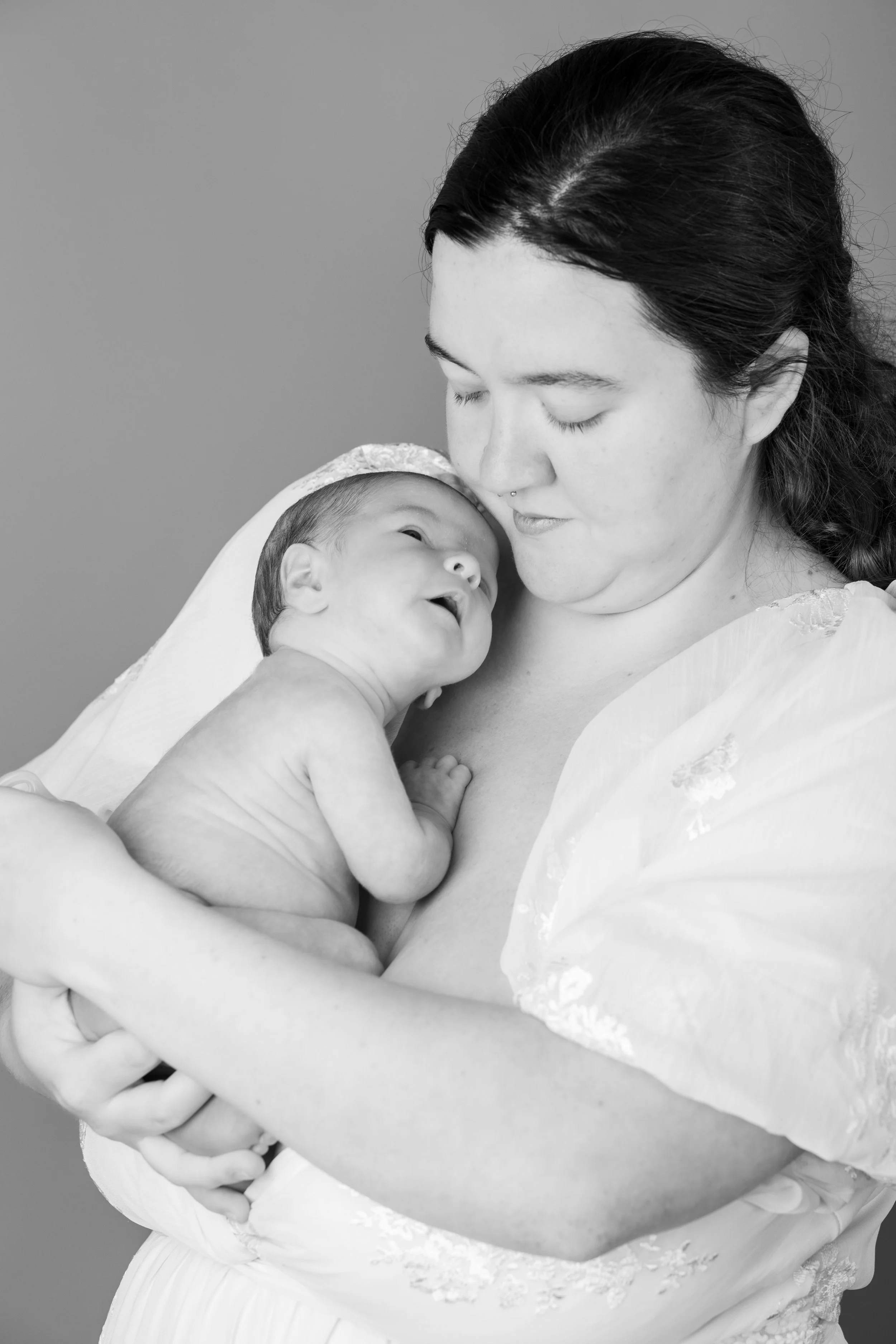Black and white photo of a woman holding a newborn baby against her chest. The woman is looking down at the baby with a gentle expression, and the baby is looking up at her, appearing happy.