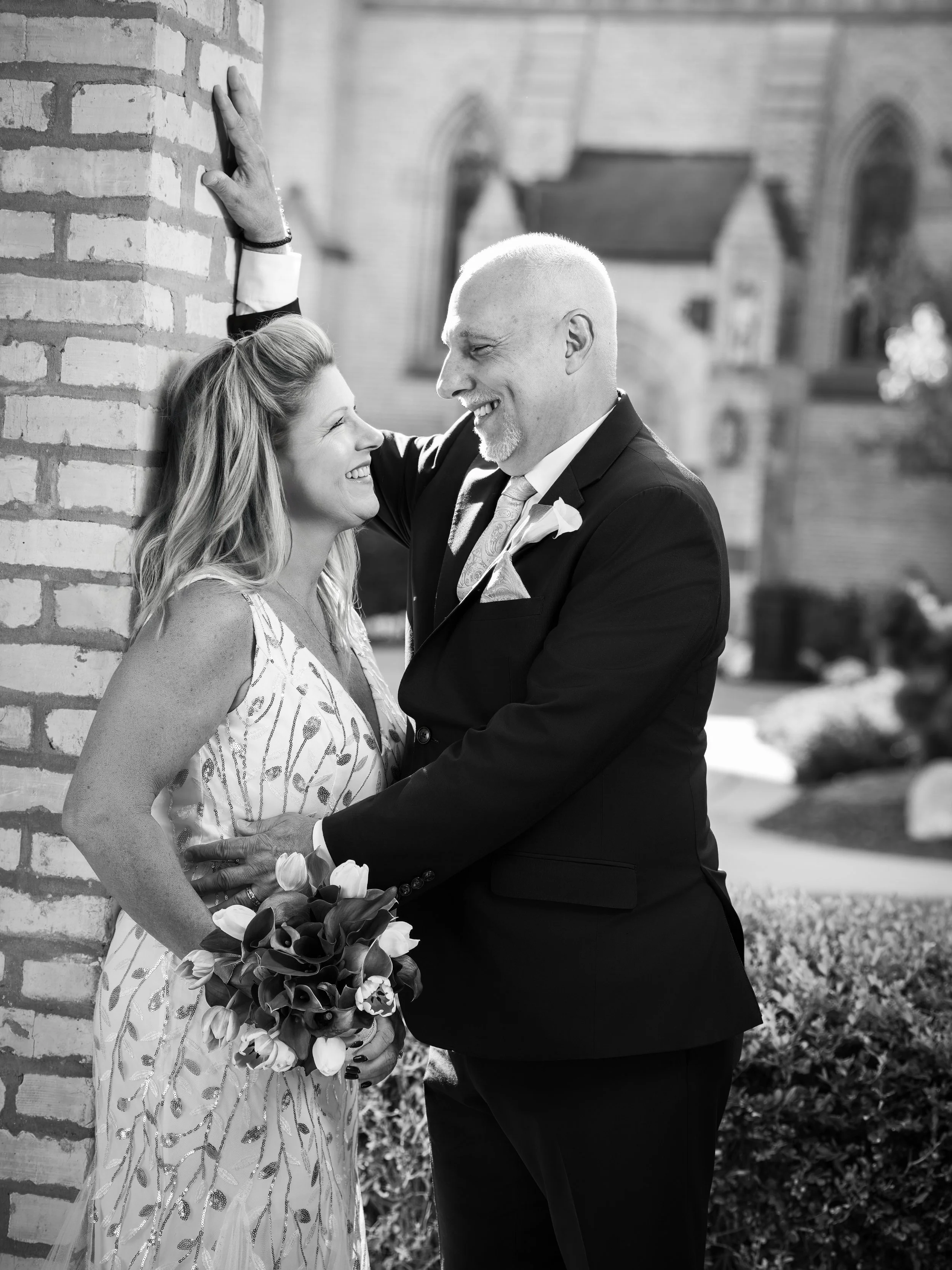 A joyous couple, the woman holding a bouquet of flowers, sharing a happy moment outside a building, with the man leaning against a brick wall.