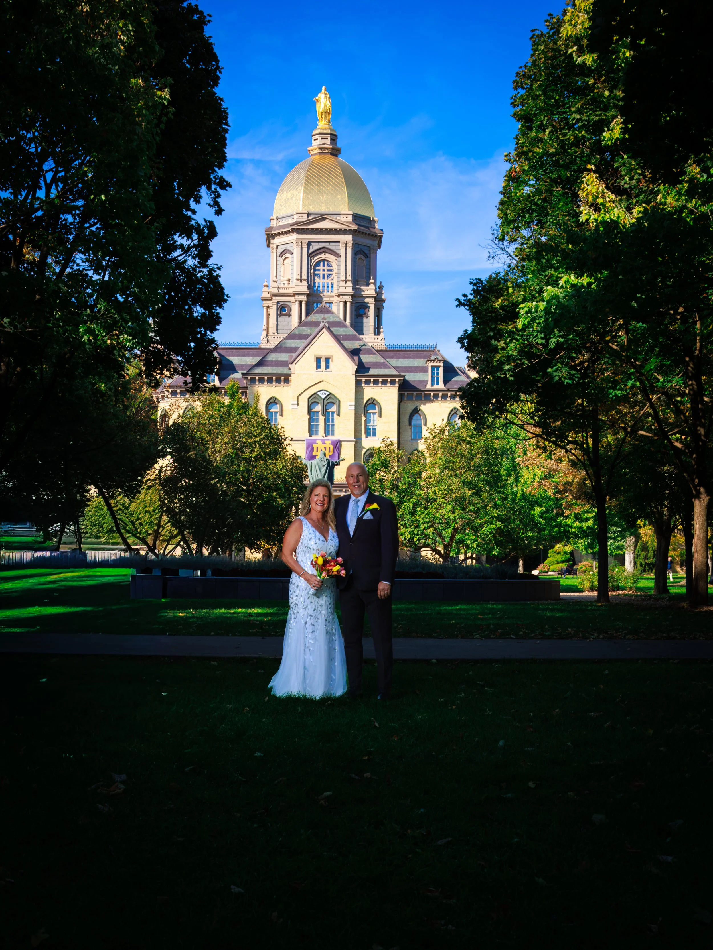 Couple in wedding attire standing on a grassy area in front of a historic building with a gold dome, trees framing the scene, and clear blue sky overhead.