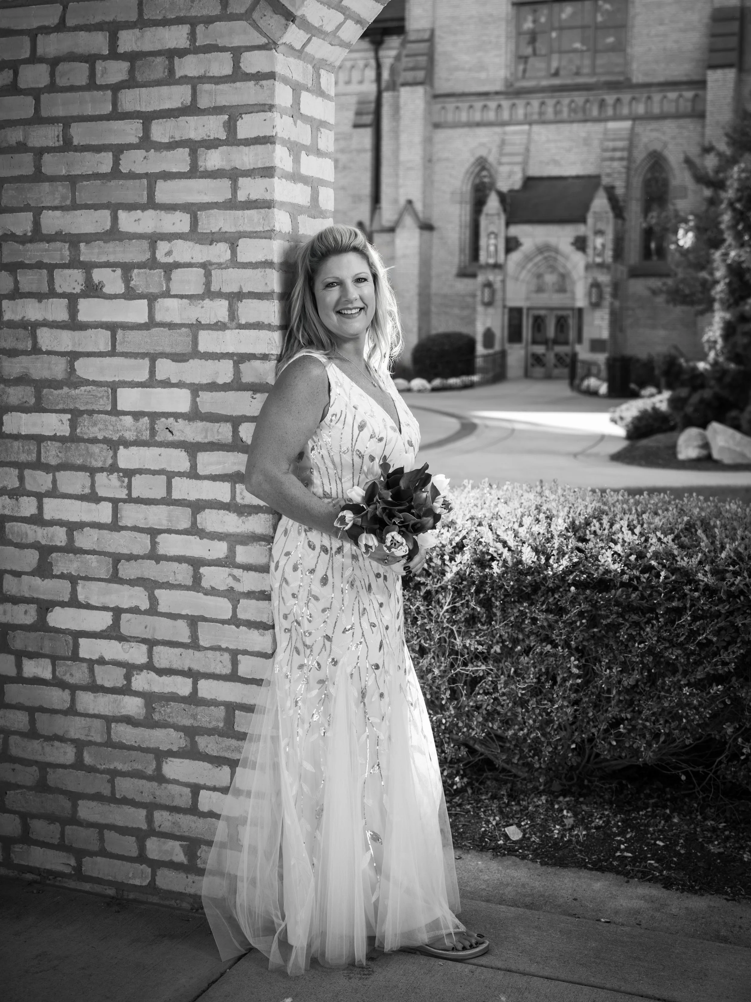 A woman in a sleeveless dress holding a bouquet of flowers, standing next to a brick wall with a church or cathedral in the background.