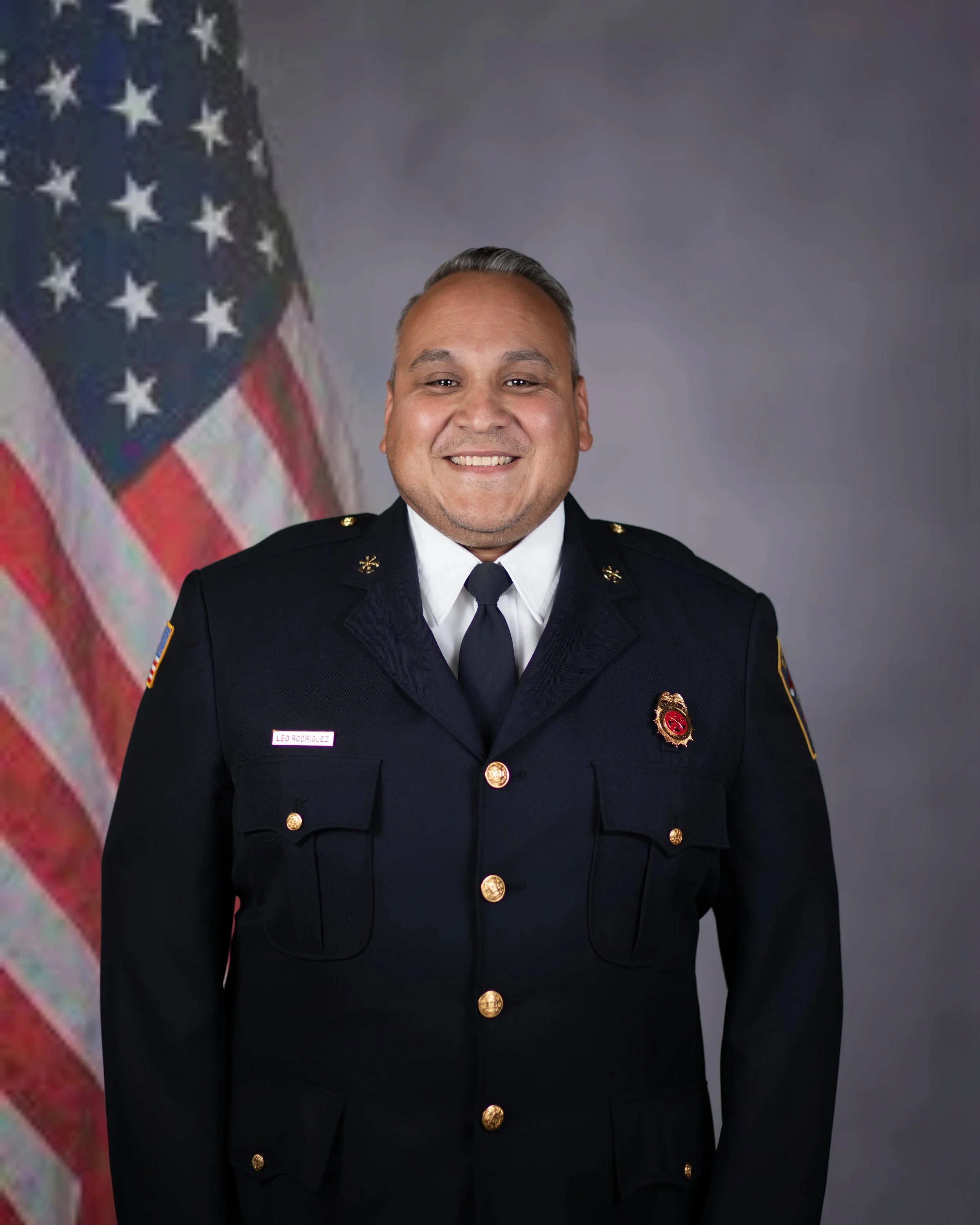A portrait of a man in a formal firefighter uniform standing in front of an American flag background.