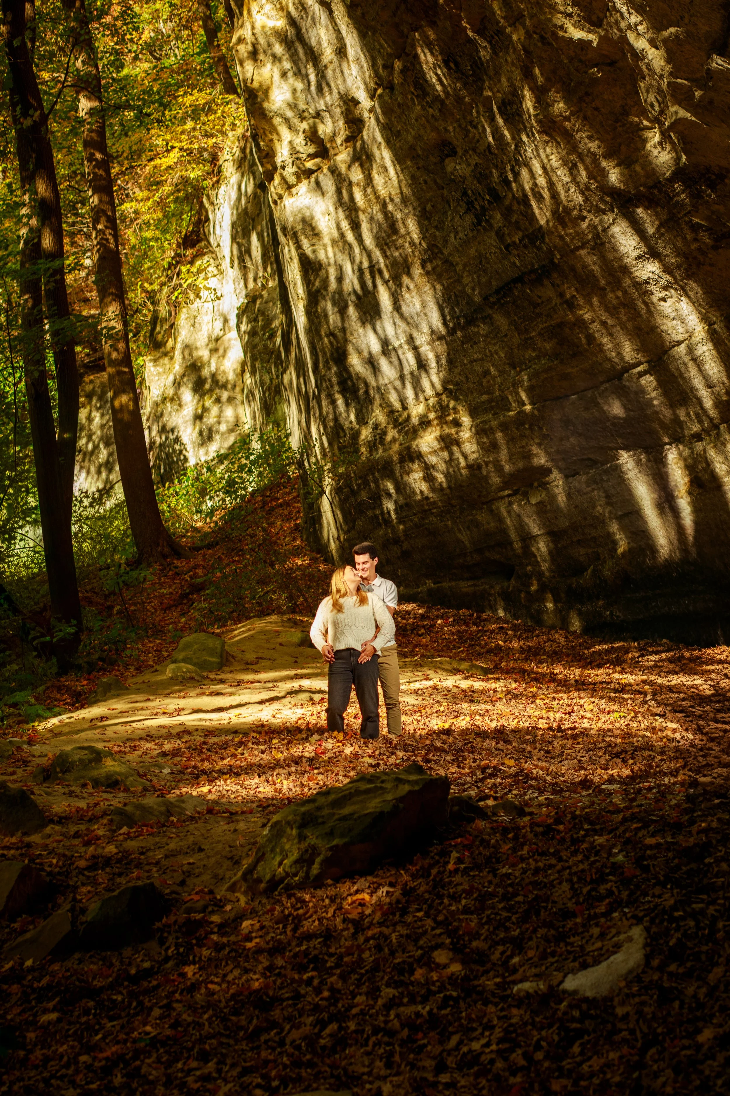A couple hugging while walking through a forest with tall trees and a rocky overhang, sunlight filtering through the leaves, and fallen leaves on the ground.