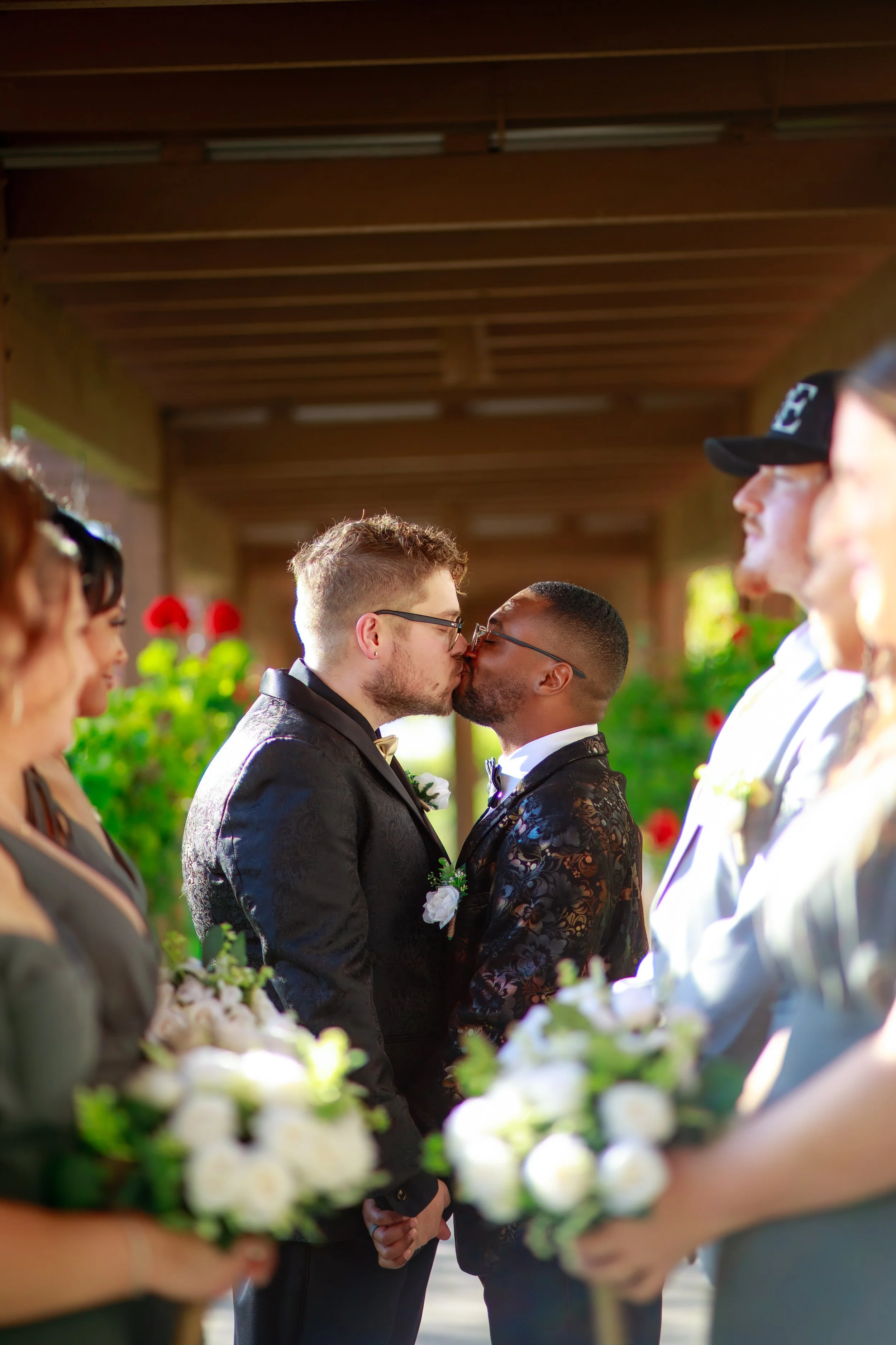 Two men in tuxedos share a kiss at a wedding ceremony while holding hands, surrounded by bridesmaids and groomsmen, with blurred greenery and flowers in the background.