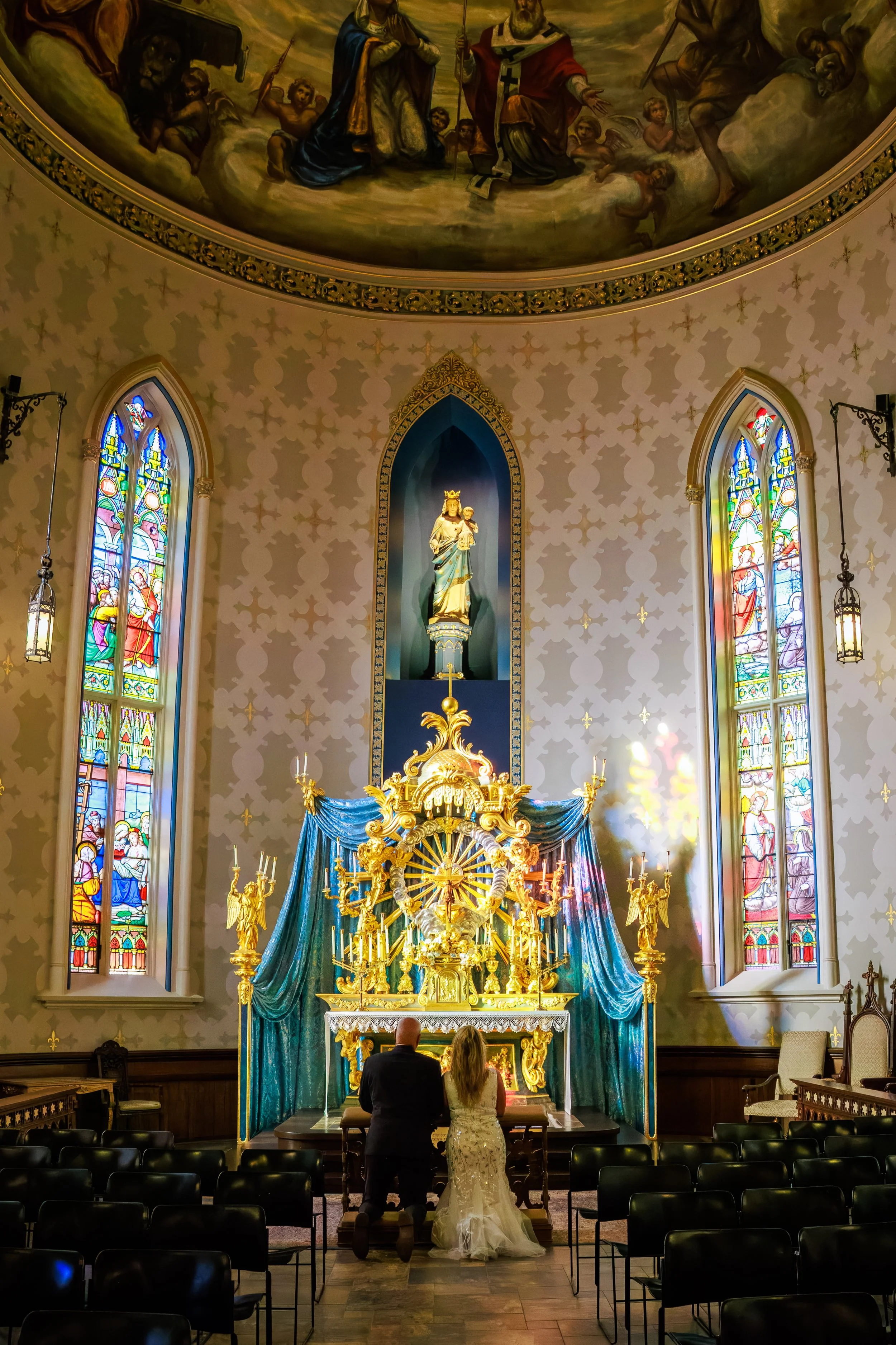 A bride and groom kneeling in prayer at the altar of a decorated church with stained glass windows, golden statues, and an ornate painting on the ceiling.
