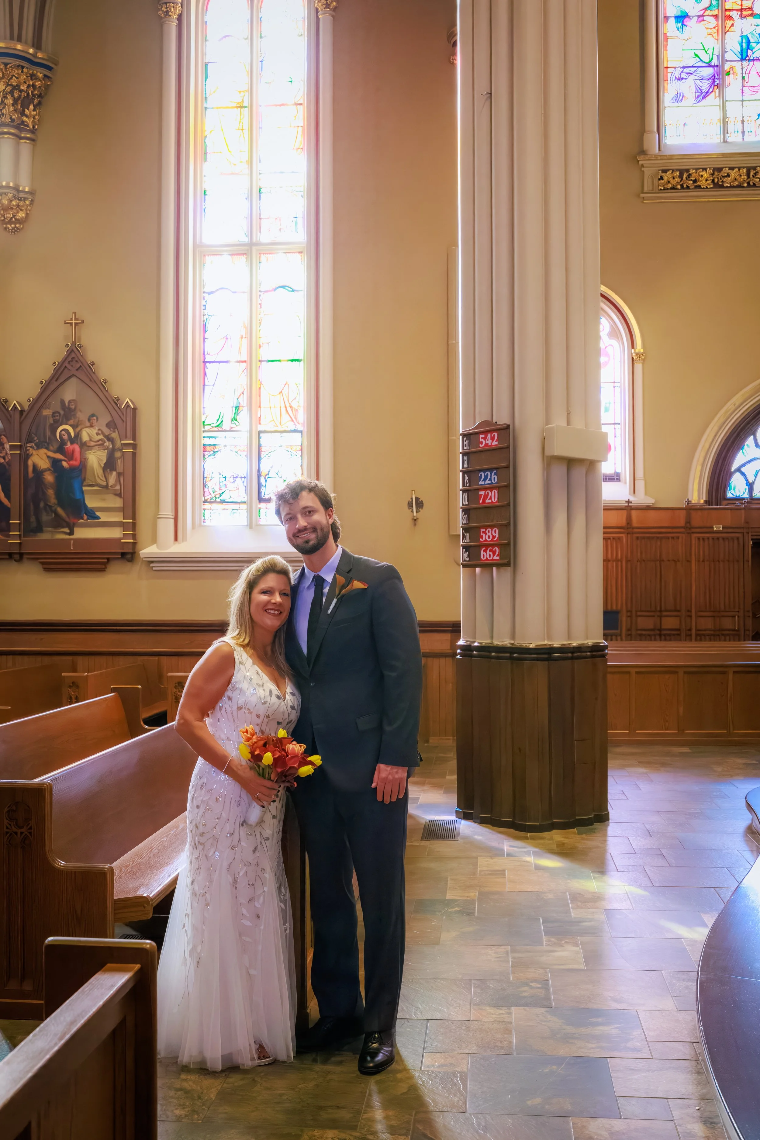 A smiling bride and groom standing inside a church, with stained glass windows and wooden pews, the bride holding a bouquet of flowers.