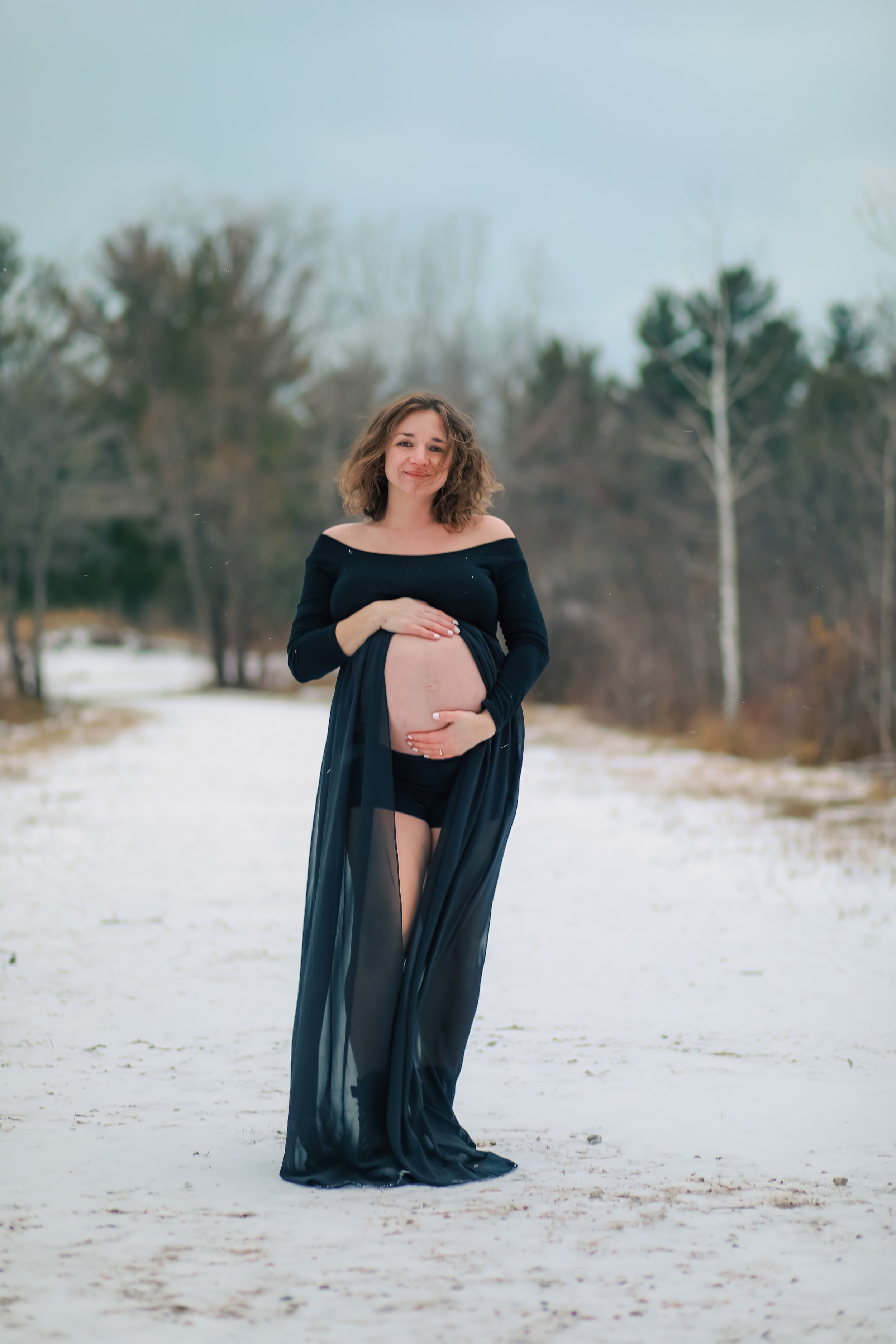 A pregnant woman standing outdoors in a snowy landscape, wearing a black off-shoulder dress with a sheer overlay, smiling gently with her left hand on her belly and her right hand on top of it.