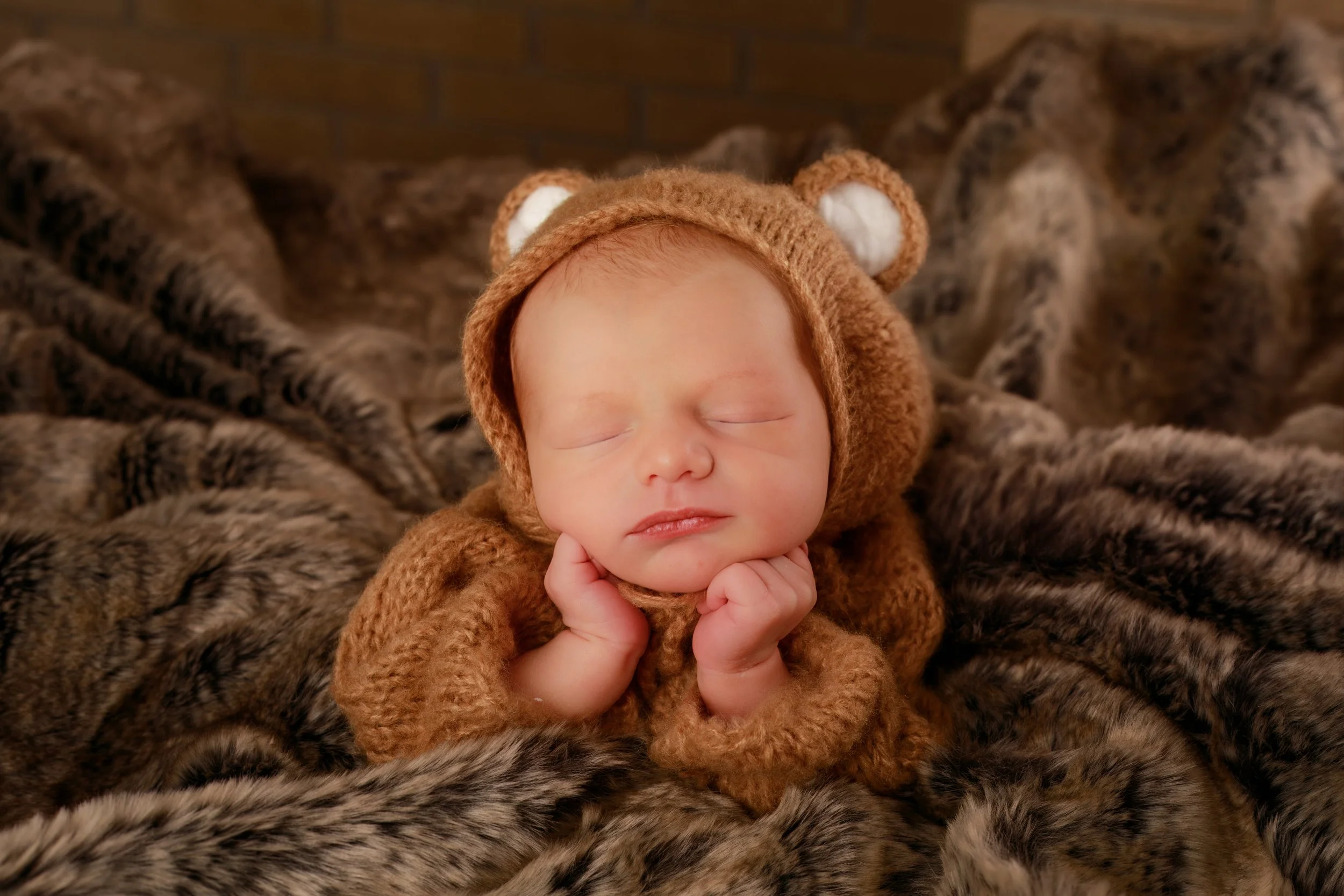 Young baby dressed in a brown bear costume, sleeping peacefully with hands under chin on a soft faux fur blanket.