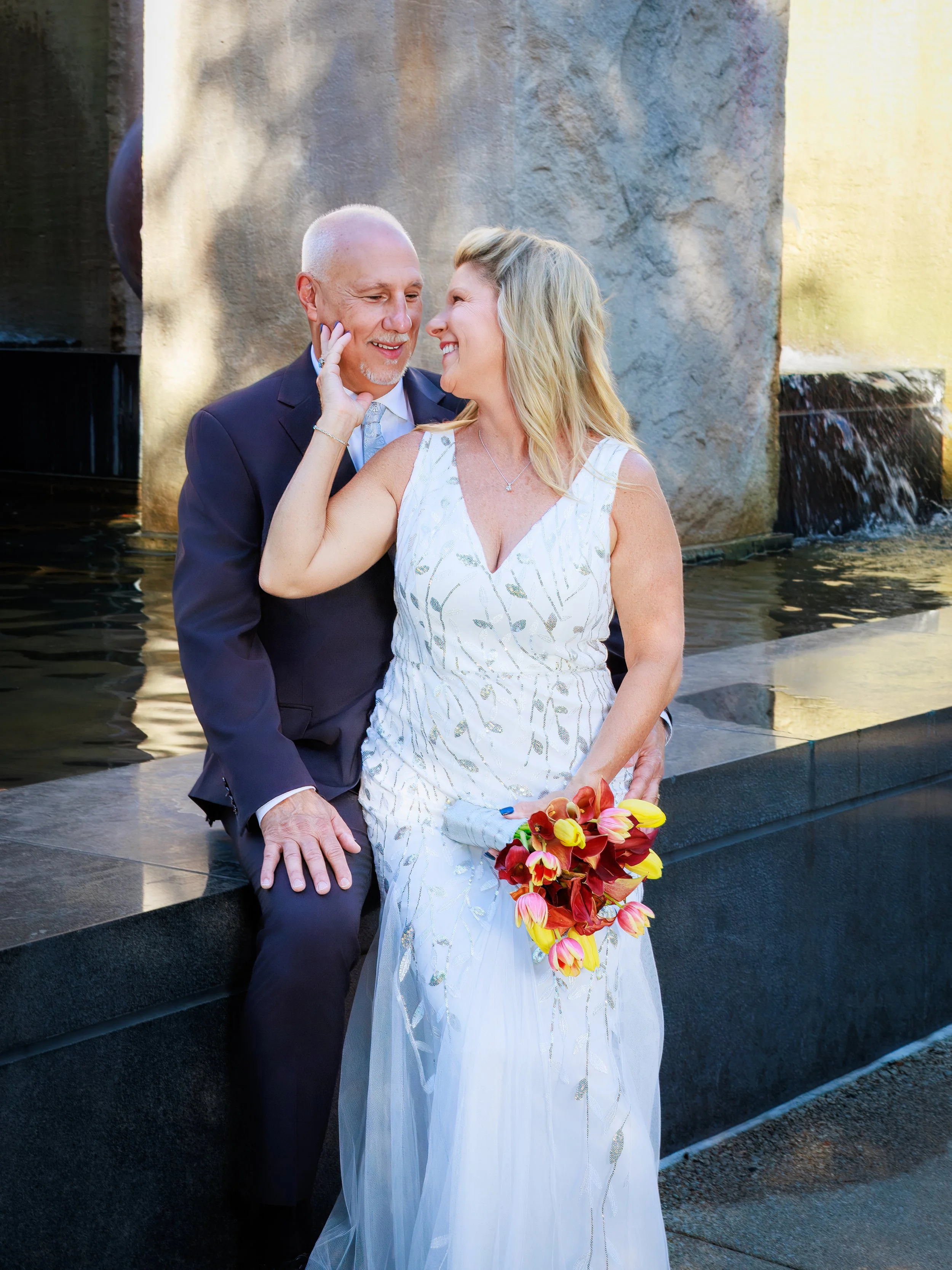 A couple dressed in wedding attire, the woman in a white dress holding a bouquet with red and yellow flowers, and the man in a dark suit, sitting close together near a water feature with a cascading fountain.