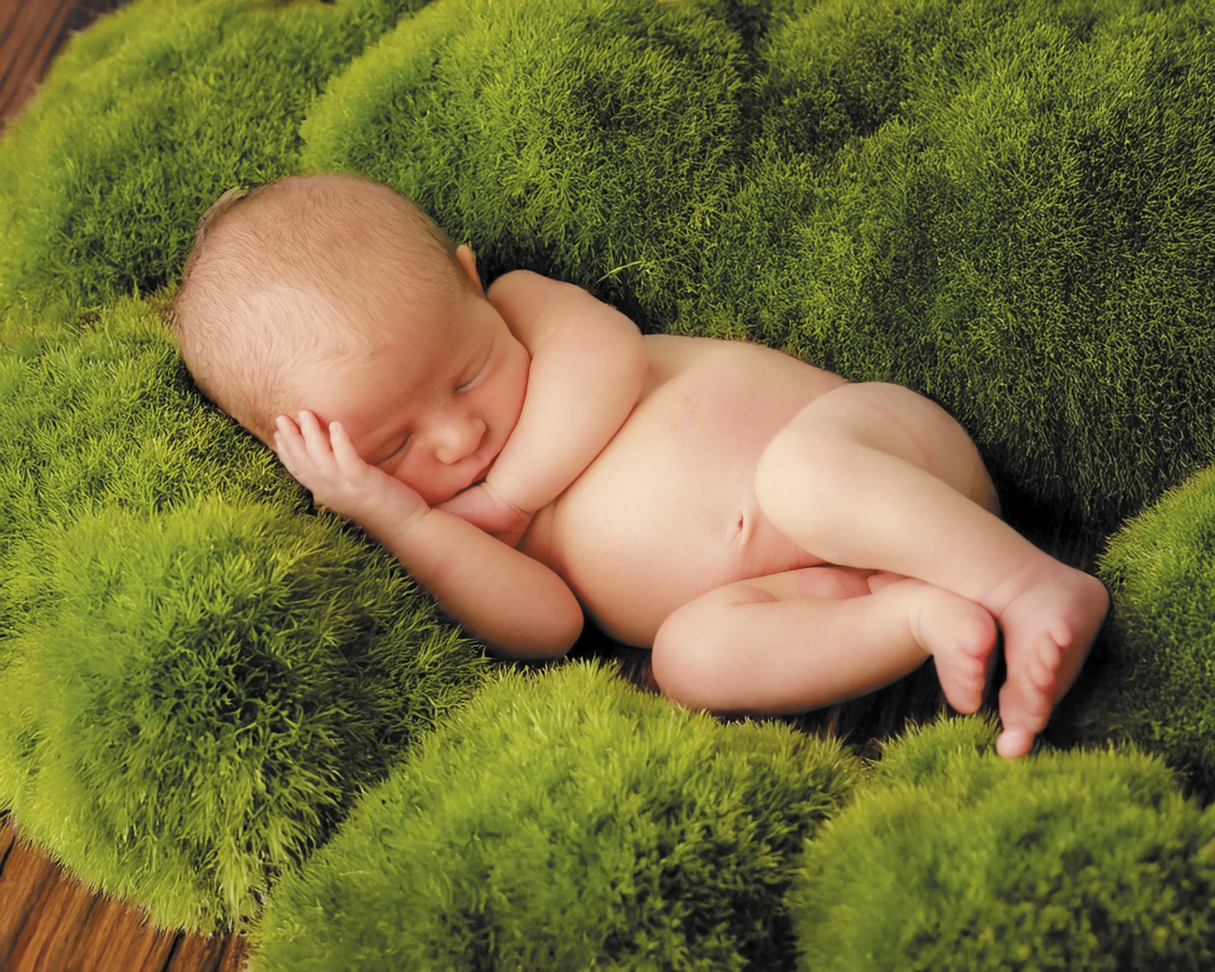 A sleeping newborn baby lying on a plush, green moss-like surface with a wooden floor visible at the edge.