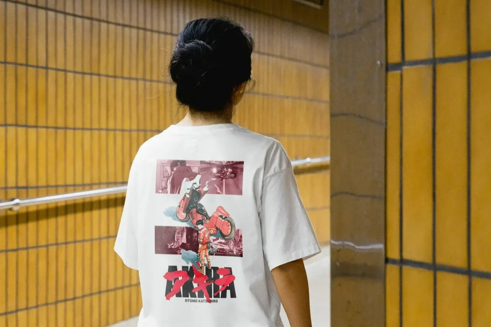 A person walking in an indoor corridor with wooden panel walls, wearing a white T-shirt featuring a graphic design with illustrations of a motorcycle, a person, and Japanese characters.