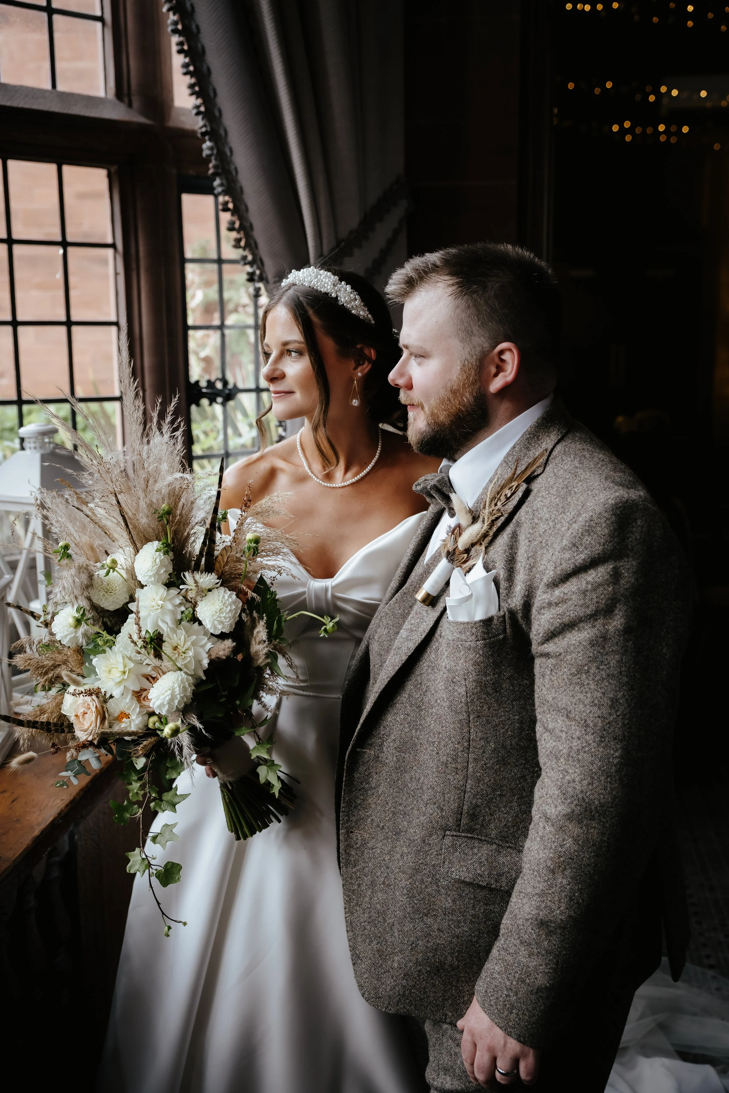 A bride and groom standing indoors near a window with curtains, dressed in wedding attire, looking out. The bride holds a large bouquet of white flowers and pampas grass, featuring lilies, roses, and greenery. The bride wears a pearl necklace and a b