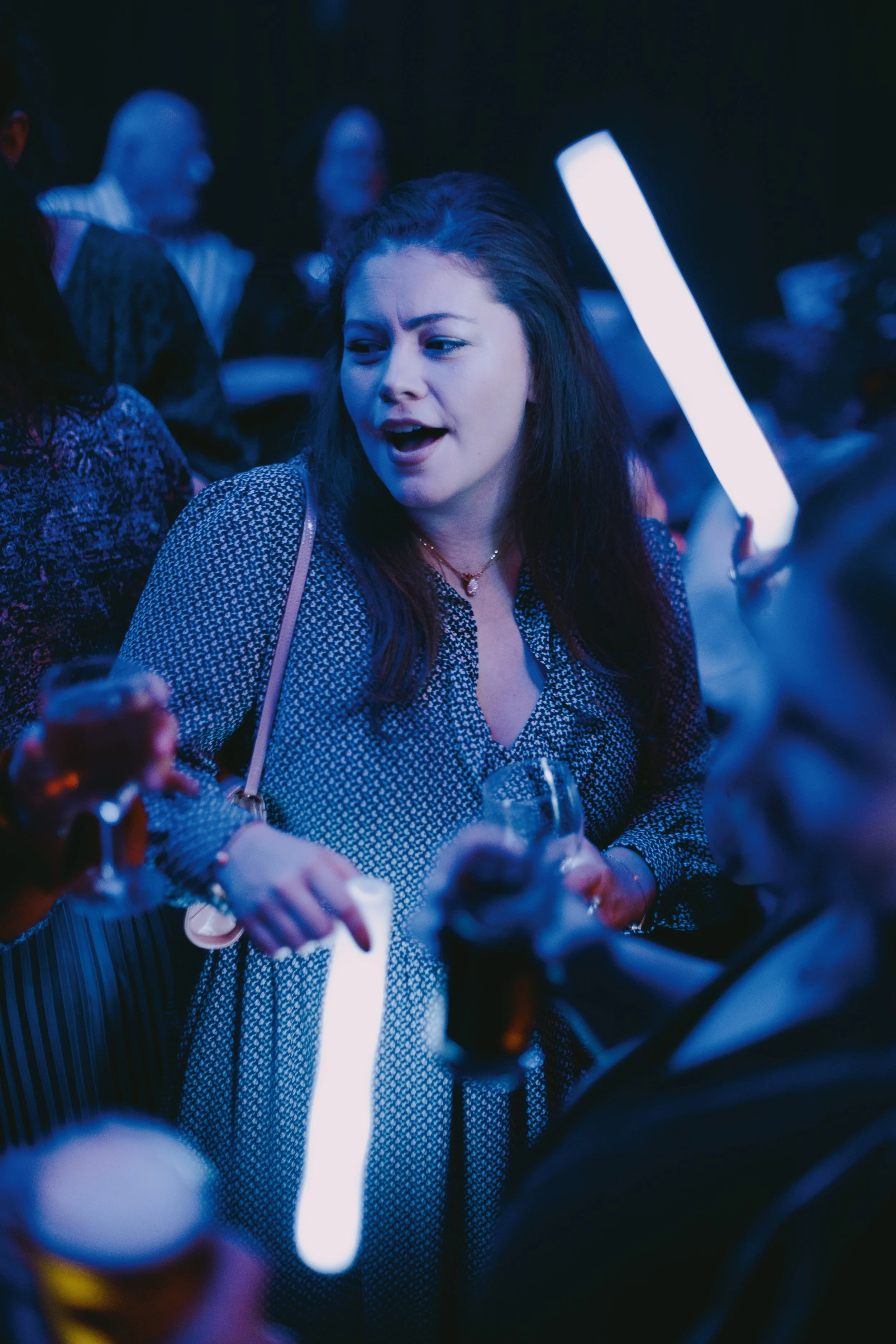 Young woman with dark hair, wearing a black and white patterned dress, holding a glass of drink, talking and engaging with others at a lively social gathering lit with blue lighting.