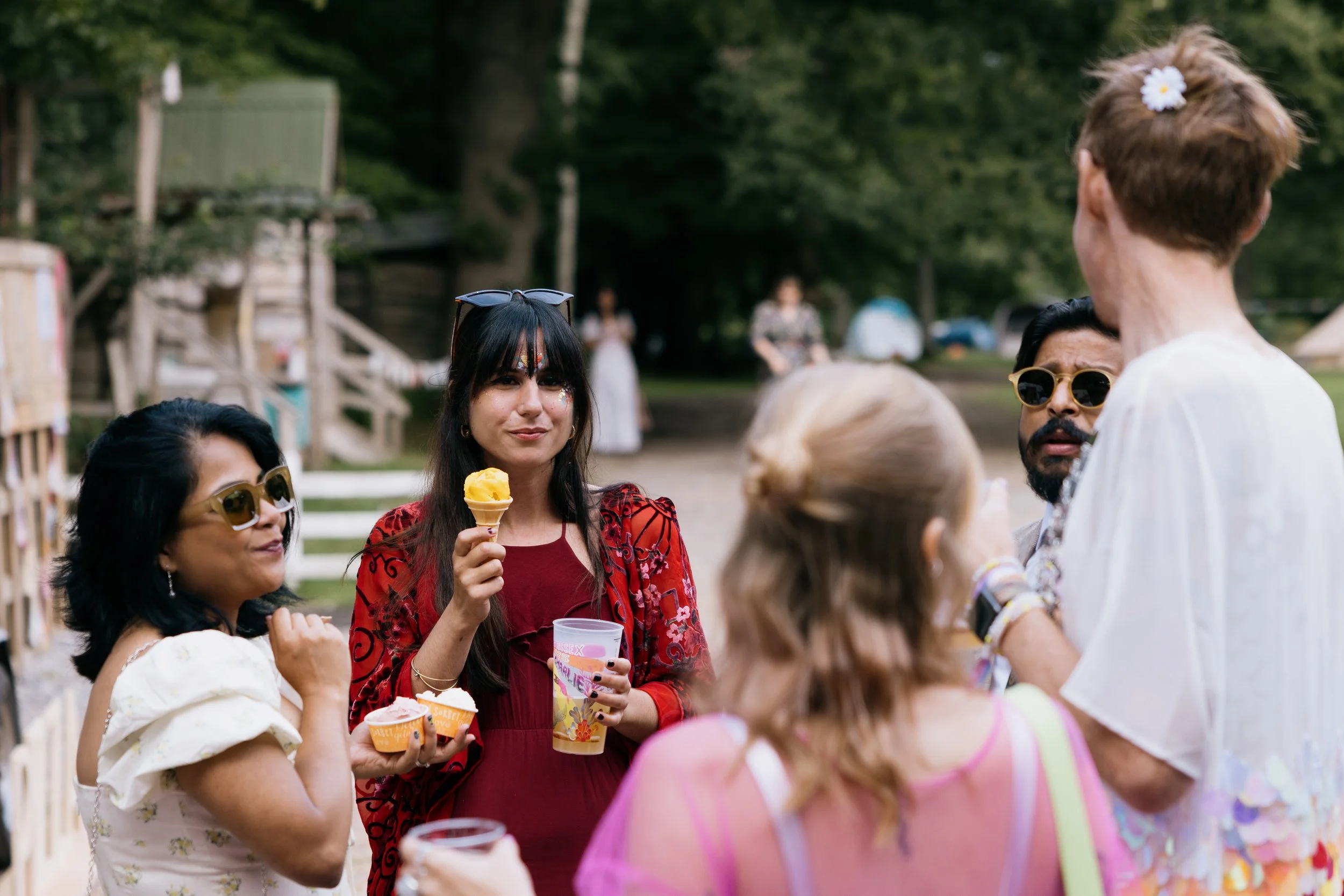 Group of friends at an outdoor summer gathering, some wearing sunglasses, holding ice cream and cupcakes, engaged in conversation, with tents and trees in the background.