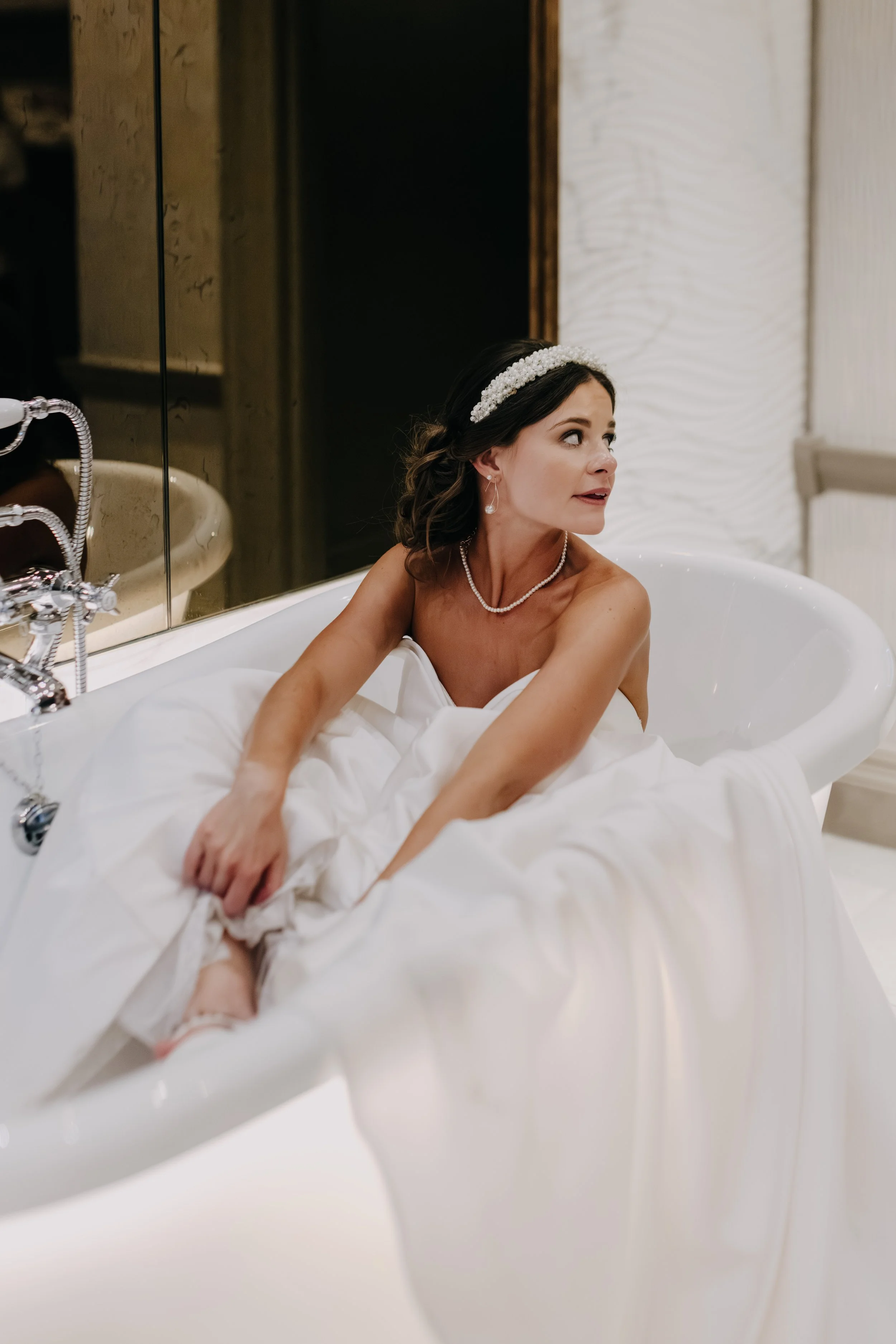 A woman in a white strapless wedding dress, wearing a pearl headband, necklace, and earrings, sitting in a bathtub with her arms resting on the edge, looking to her side in a room with elegant decor.